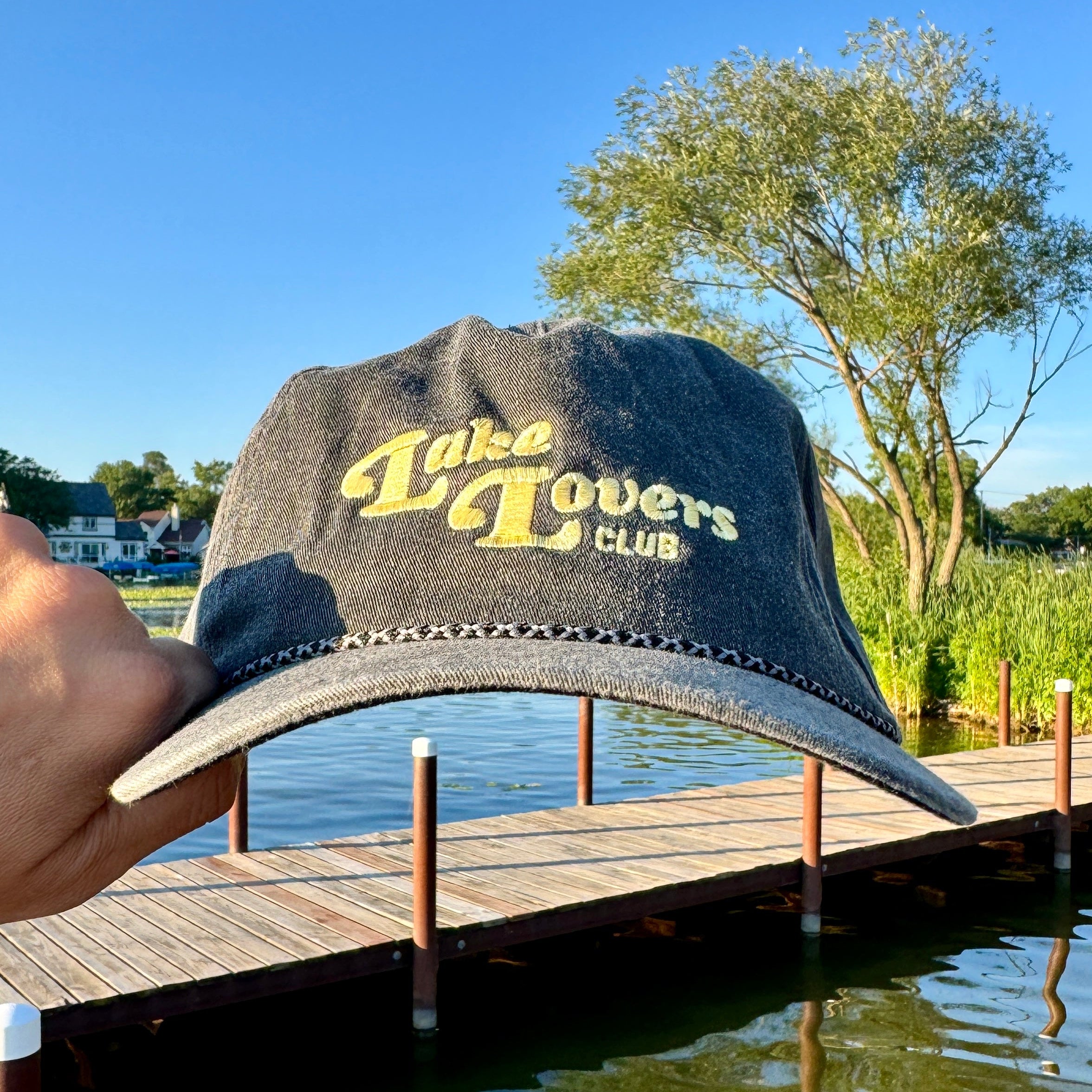 A hand holds a dark gray washed denim baseball cap with yellow embroidered text reading 'Lake Lovers Club' in front of a wooden dock and lake water under a blue sky.