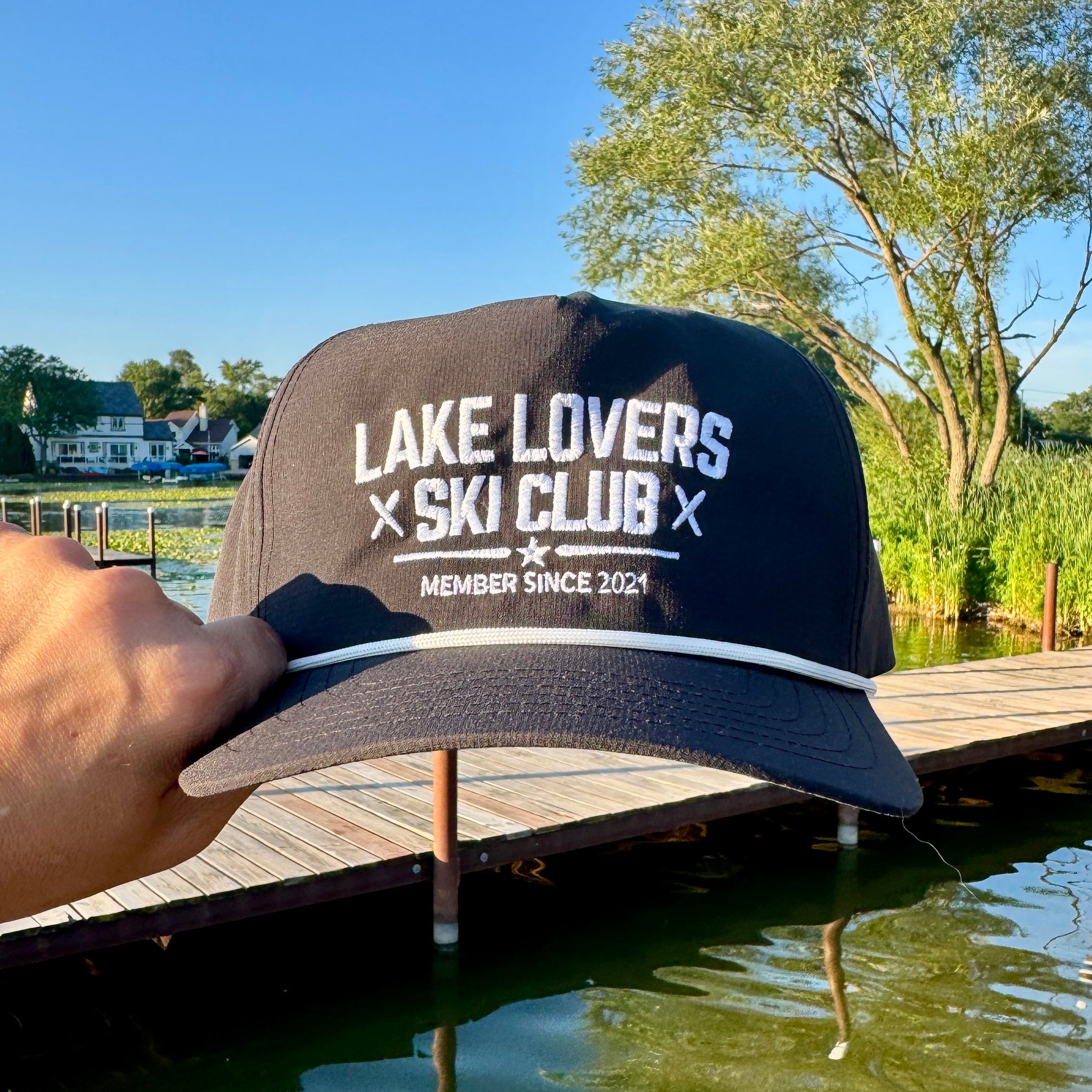 A hand holds a black snapback cap with white text reading 'LAKE LOVERS X SKI CLUB' and 'MEMBER SINCE 2021' against an outdoor lakeside background with a wooden dock, trees, and blue sky.