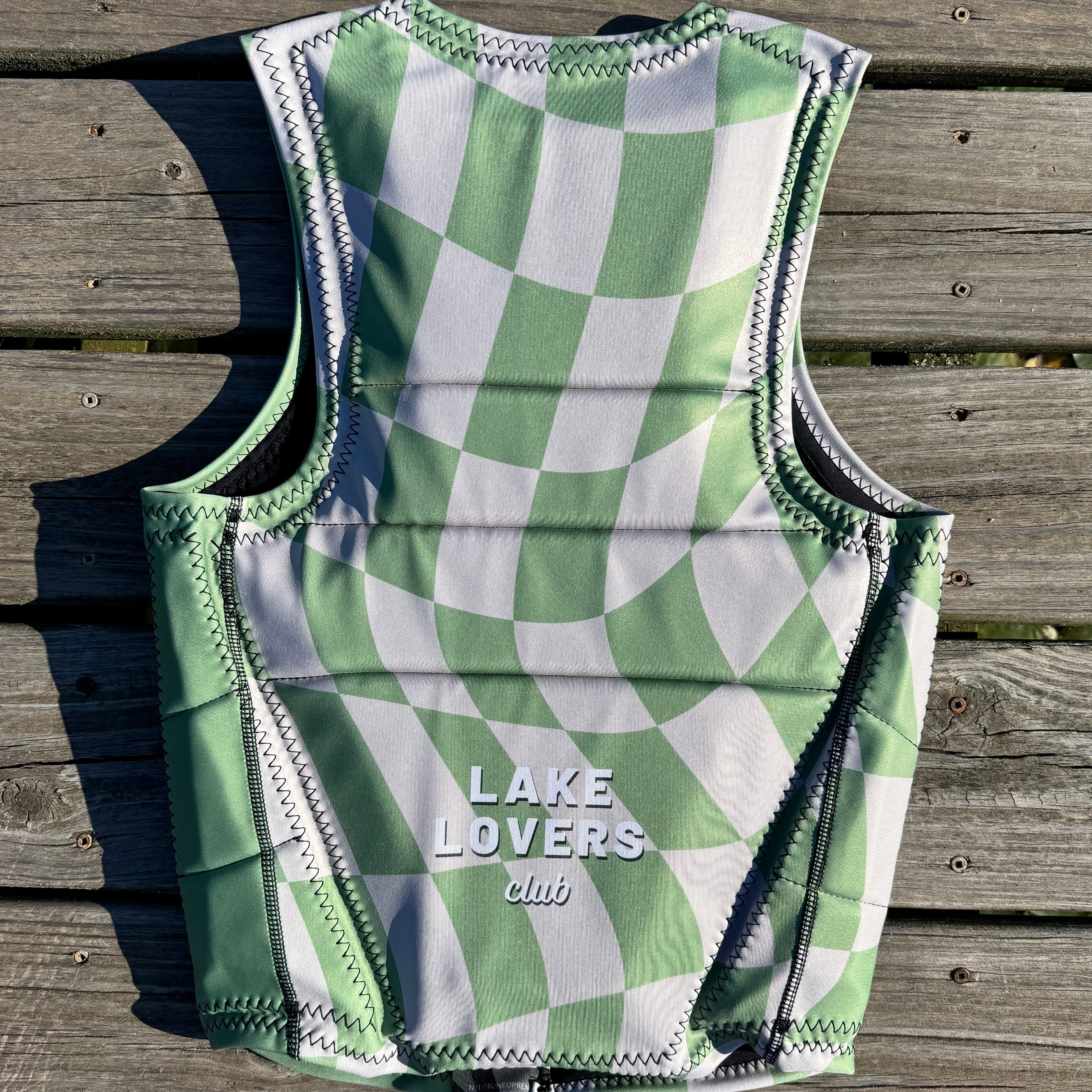 A sleeveless water sports vest with a green and white checkerboard pattern, laid flat on a weathered wooden dock. The vest features the text 'LAKE LOVERS club' printed in white near the bottom center.