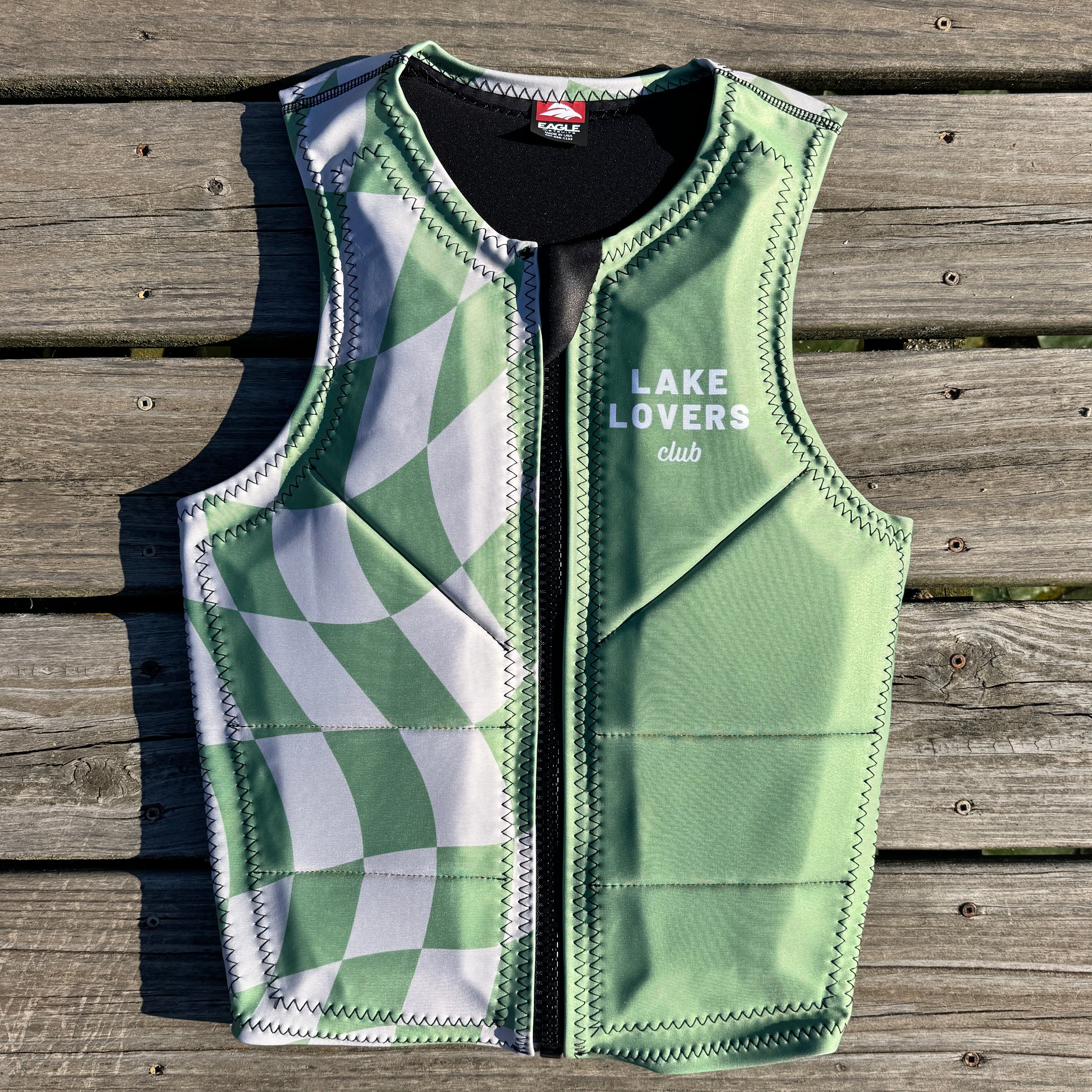 A green and white checkered water sports vest with 'LAKE LOVERS club' printed on the chest, laid flat on a weathered wooden deck.