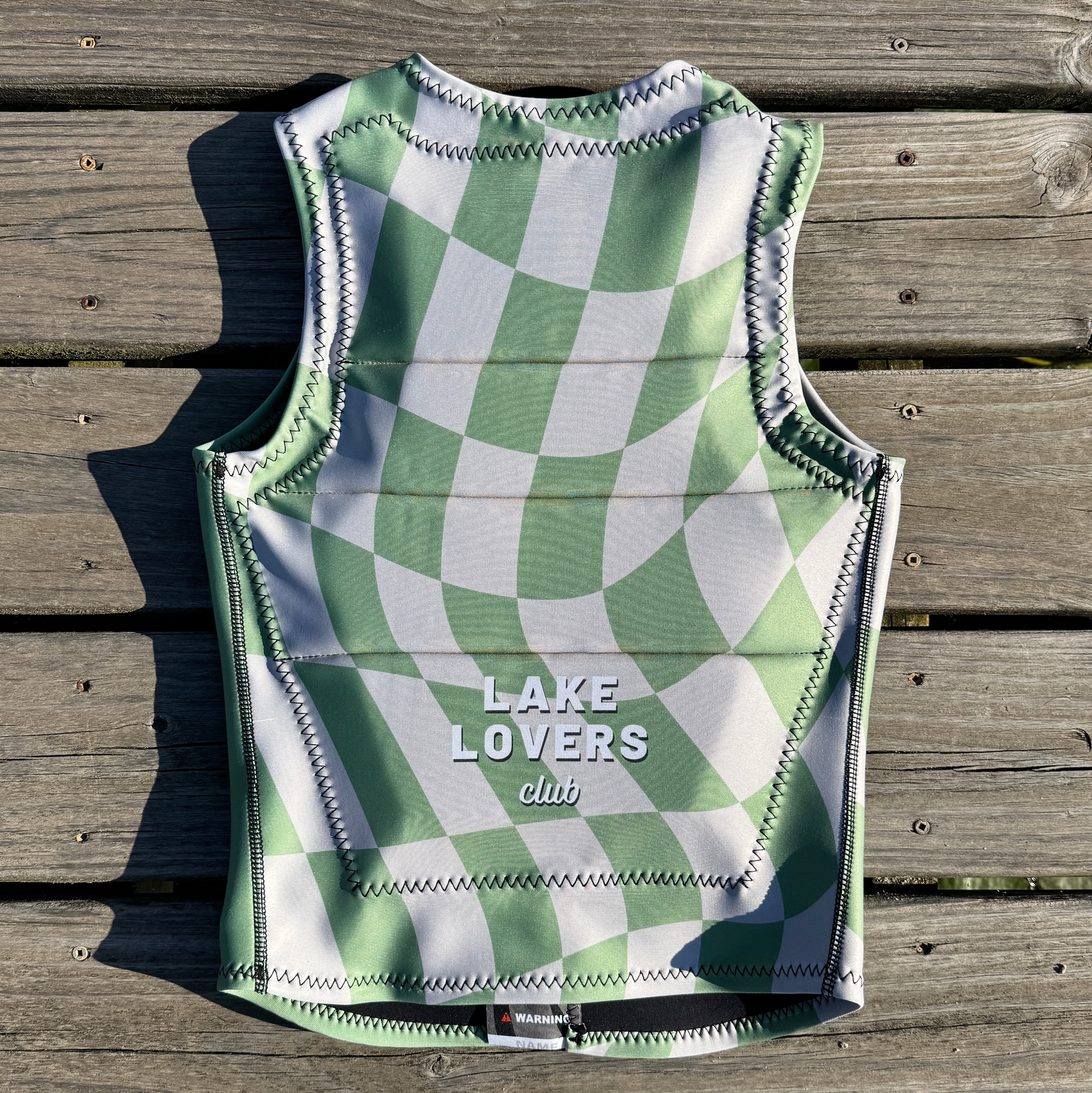 A water sports west with a green and white wavy checkerboard pattern, laid flat on a weathered wooden plank surface. The brand name 'LAKE LOVERS club' is printed in white on the lower front.