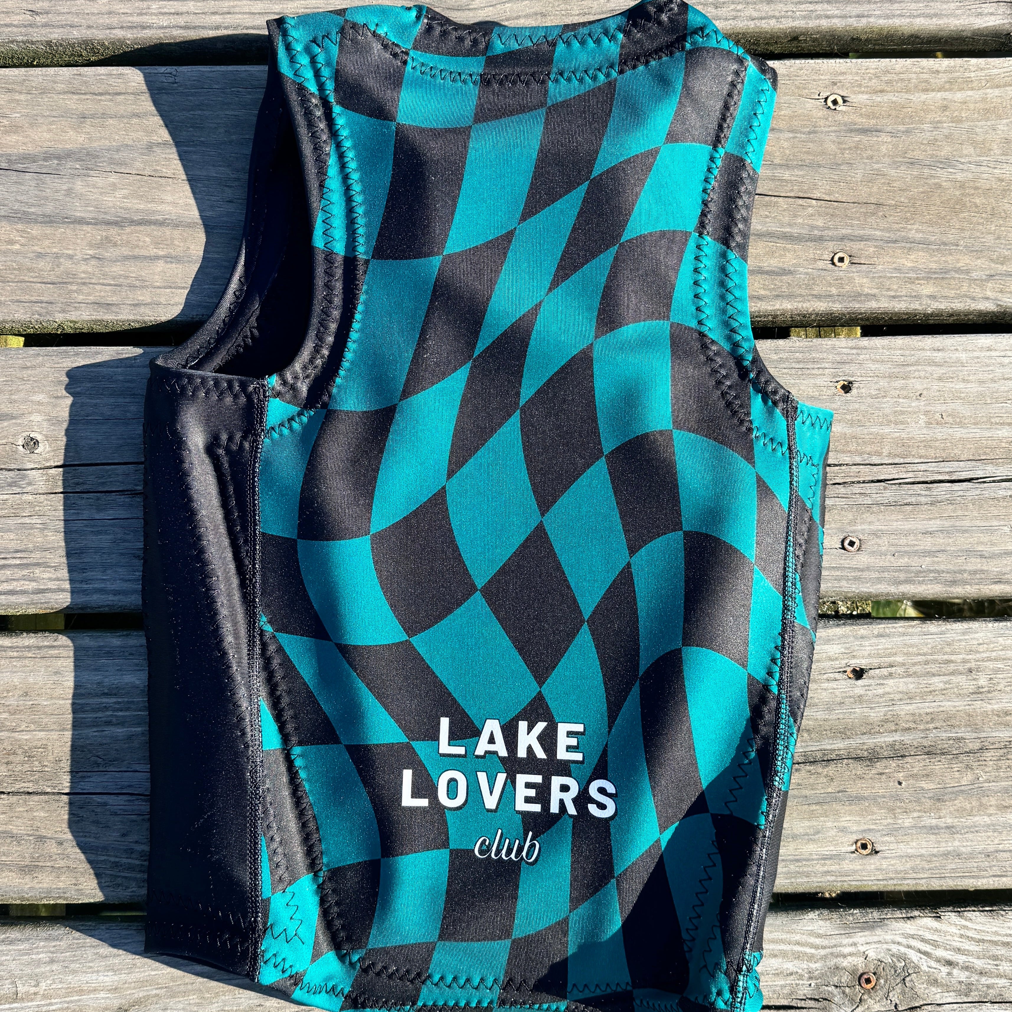 A water sports vest with a black and teal checkered pattern, featuring the white text 'LAKE LOVERS club' on the front, laid flat on a weathered wooden plank surface.