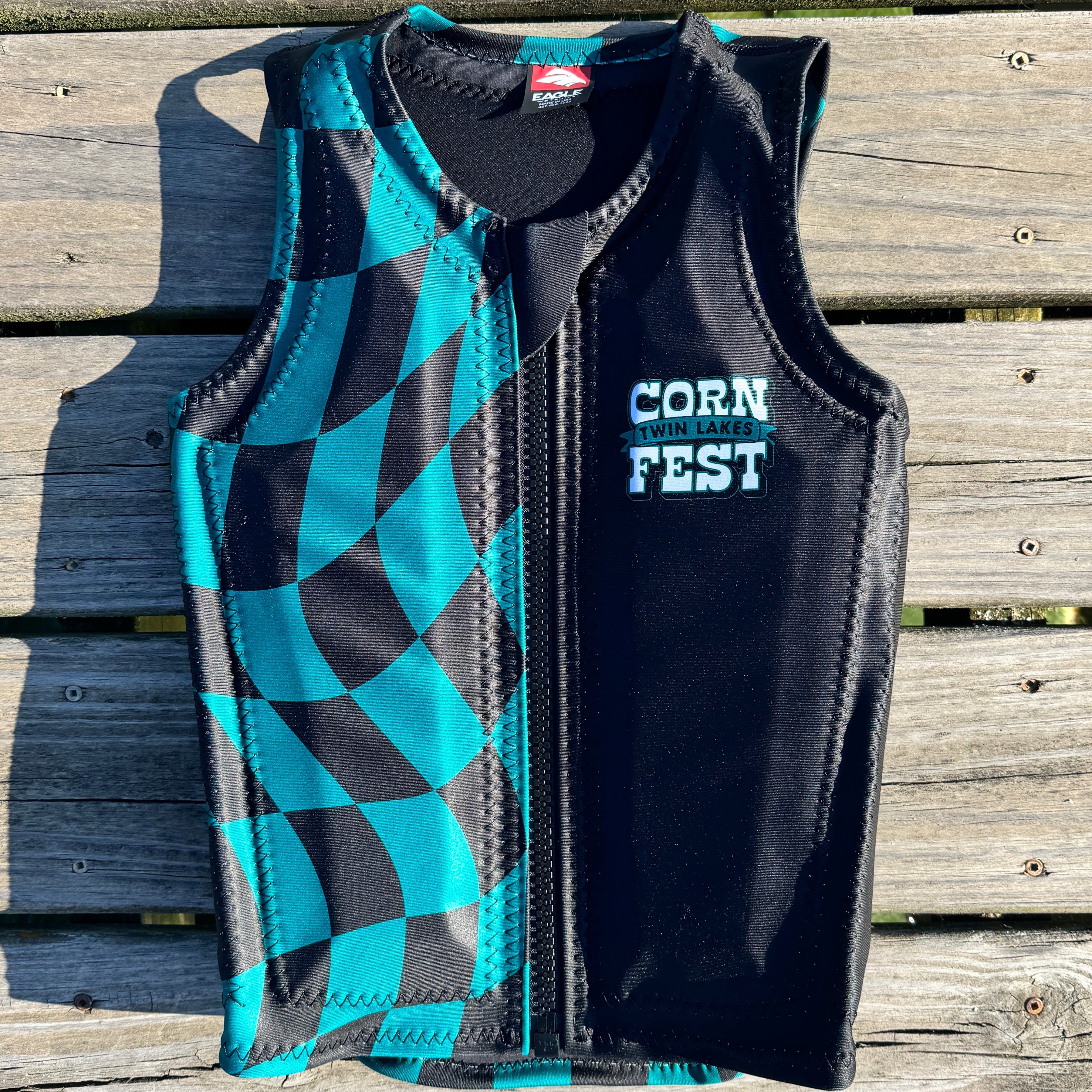 A black and teal checkered zip-up water sports vest laid flat on a weathered wooden deck. The right side of the vest is solid black with 'TWIN LAKES CORN FEST' printed in white on the chest, and the left side features a teal and black checkered pattern.