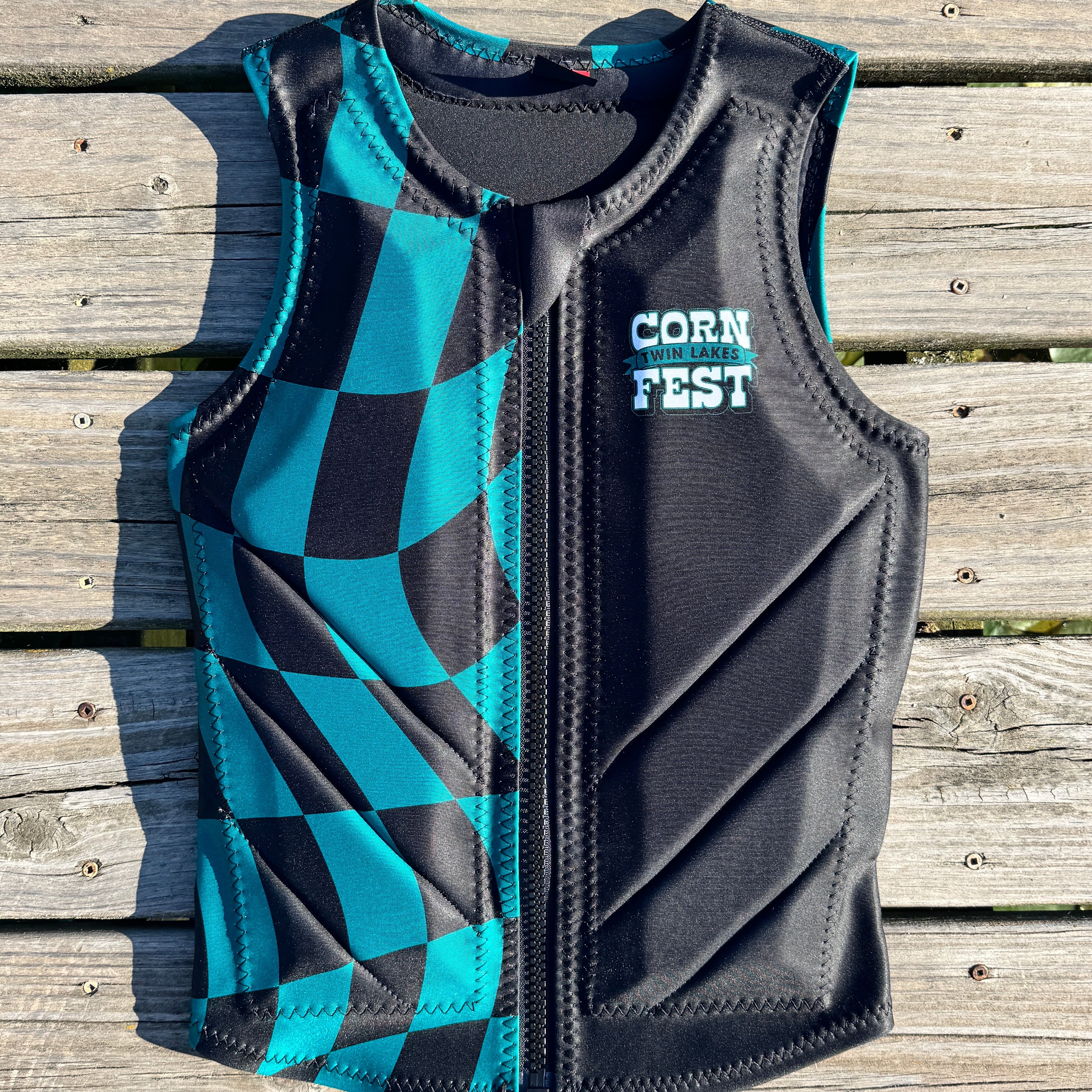 A black and teal checkered zip-up water sports vest with 'TWIN LAKES CORN FEST' printed on the chest, laid flat on a weathered wooden plank surface.