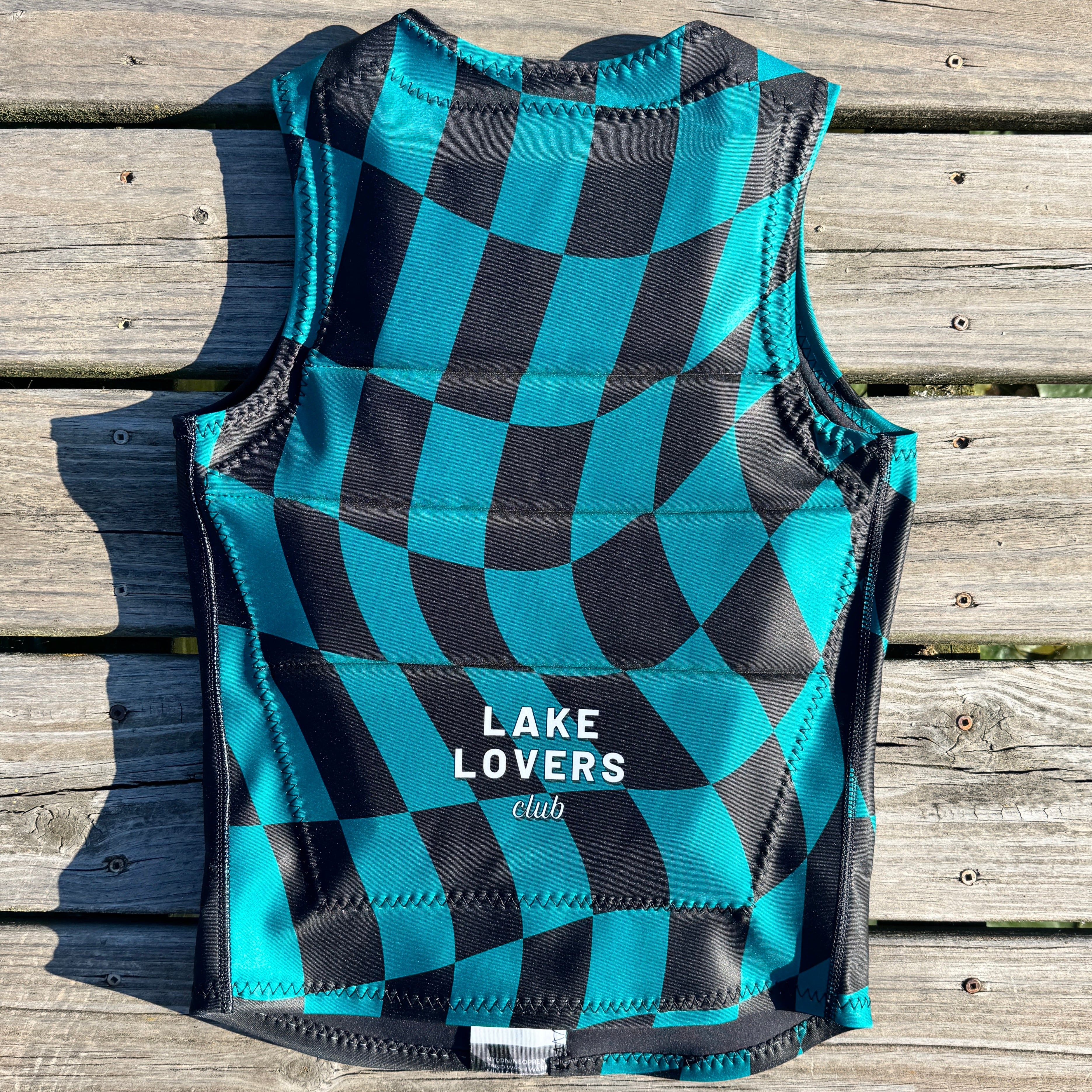 A sleeveless comp vest with a black and teal checkered pattern, laid flat on weathered wooden planks. The bottom features the white text 'LAKE LOVERS club'.