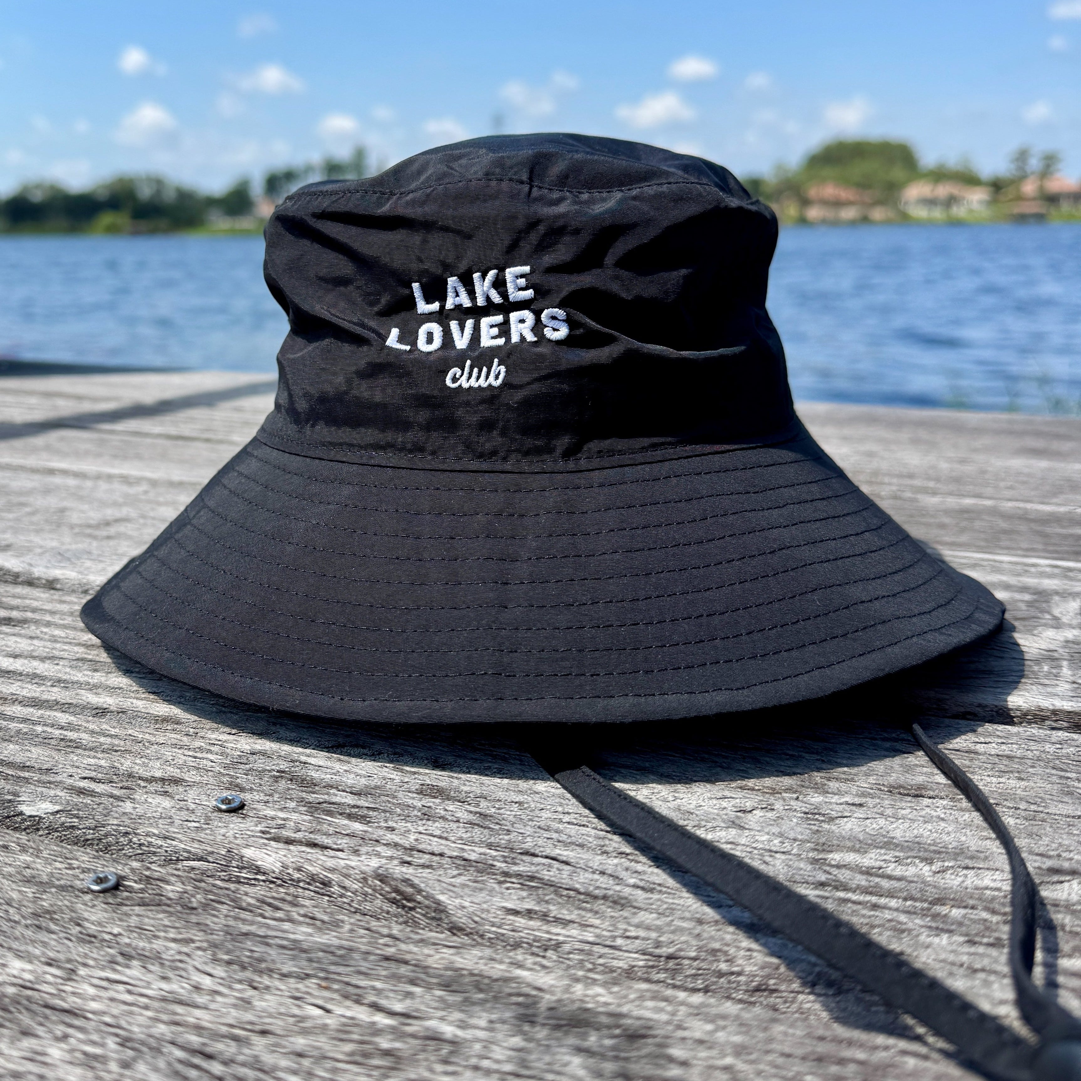 Black bucket hat with 'Lake Lovers Club' text on a wooden dock by a lake.
