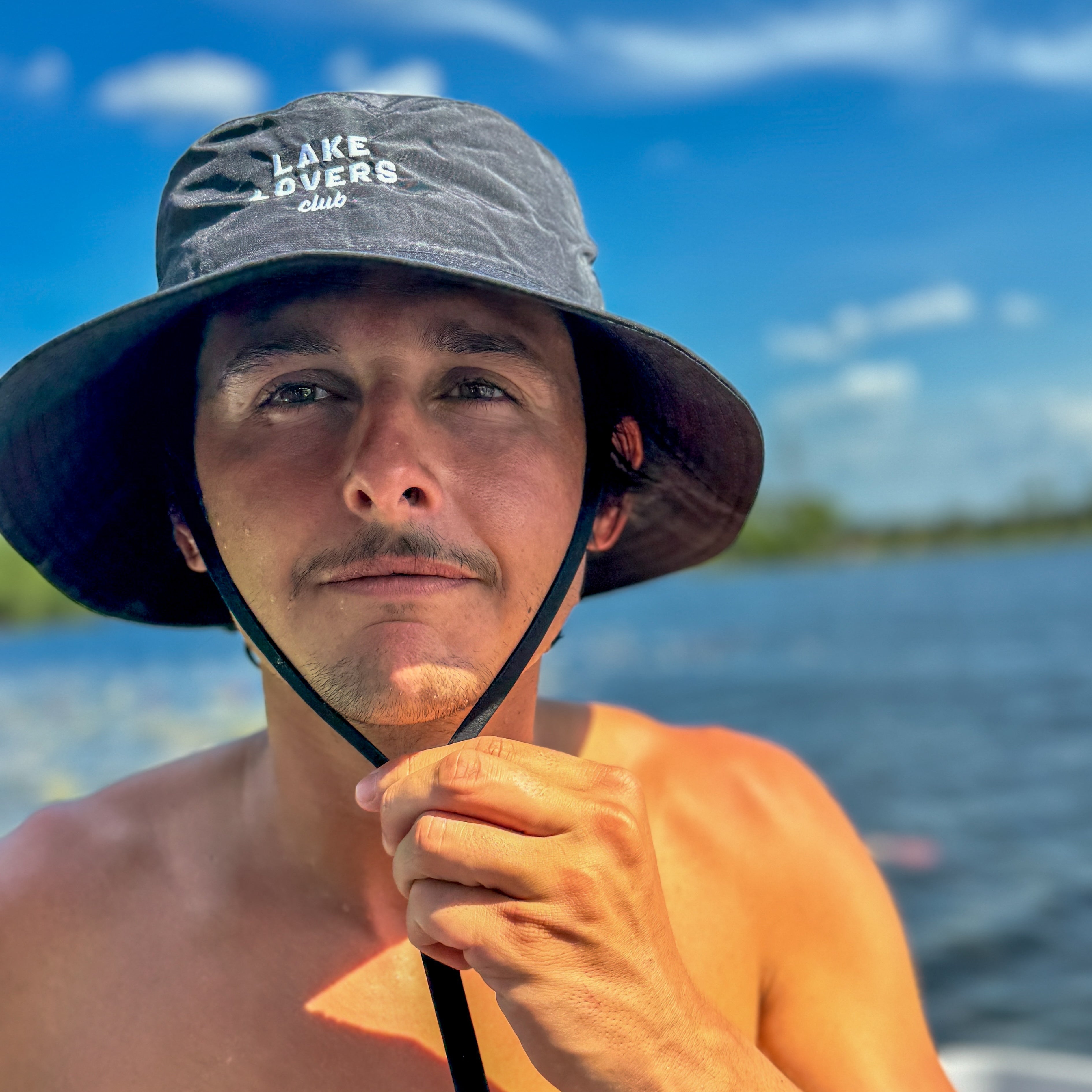 Man wearing a wide-brimmed hat with text stating 'Lake Lovers Club,' standing by a body of water under a blue sky.