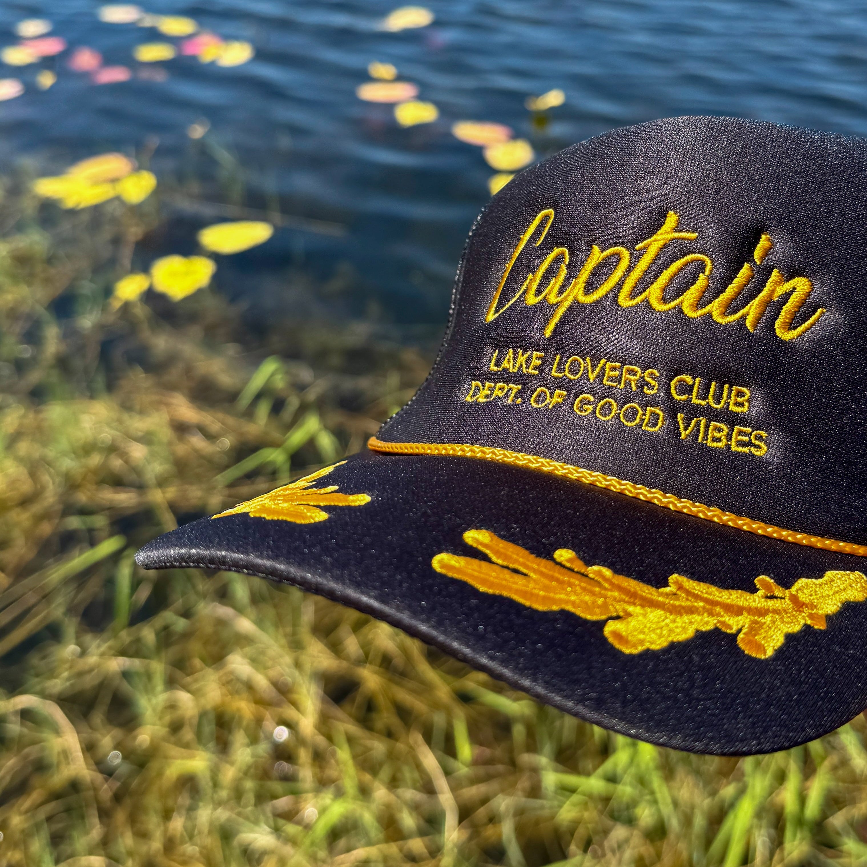 Black cap with yellow text 'Captain' and 'Lake Lovers Club Dept. of Good Vibes' against a water and grass background.