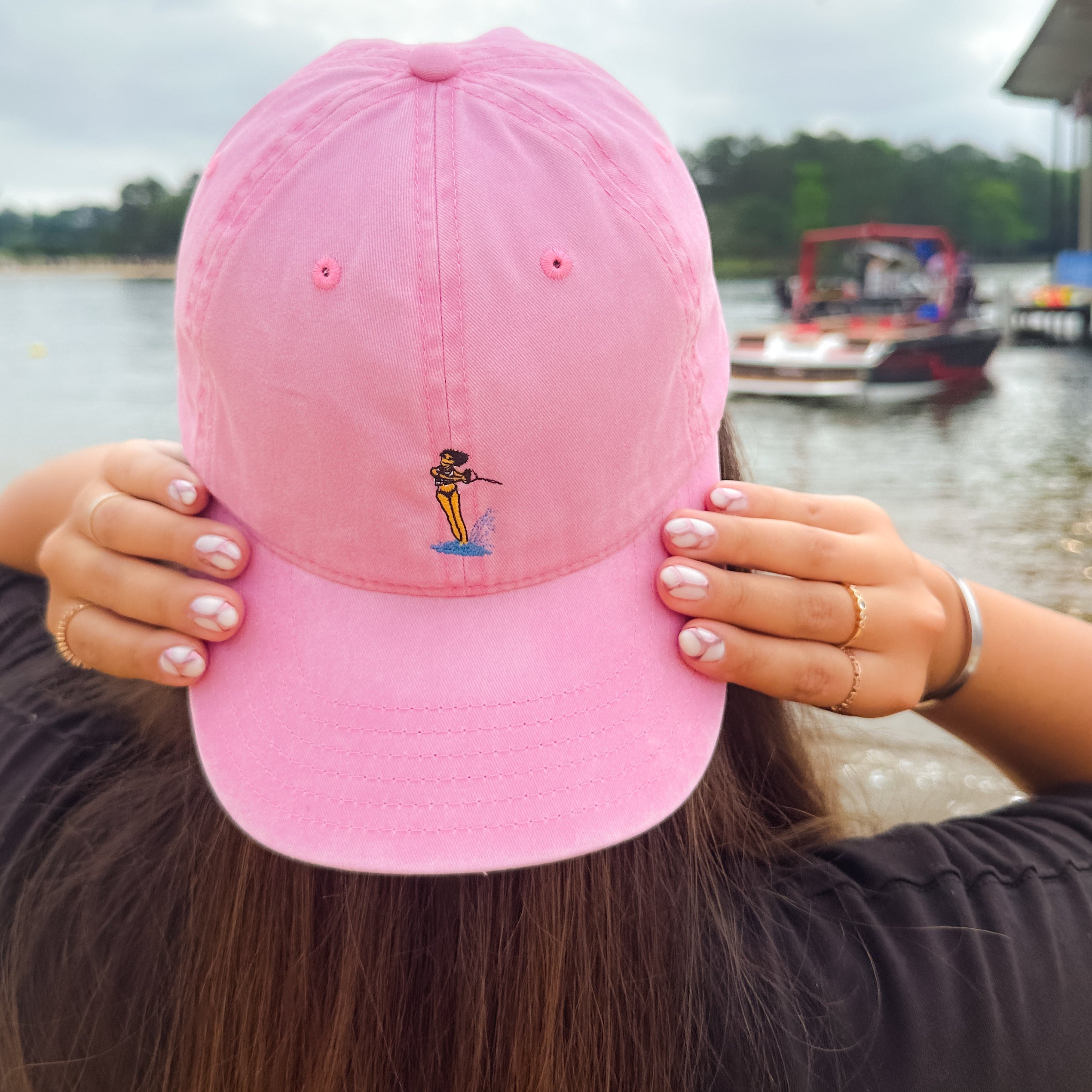 Girl with long brown hair, facing a lake and wearing a pink hat with a waterski girl design on it.