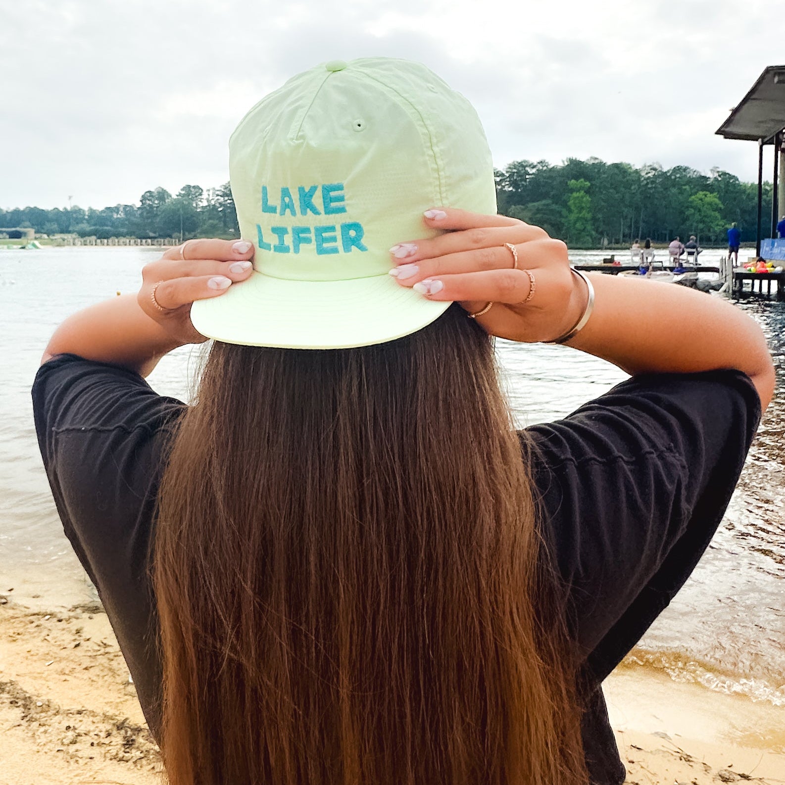 Person wearing a light lime green 'Lake Lifer' cap by a lake