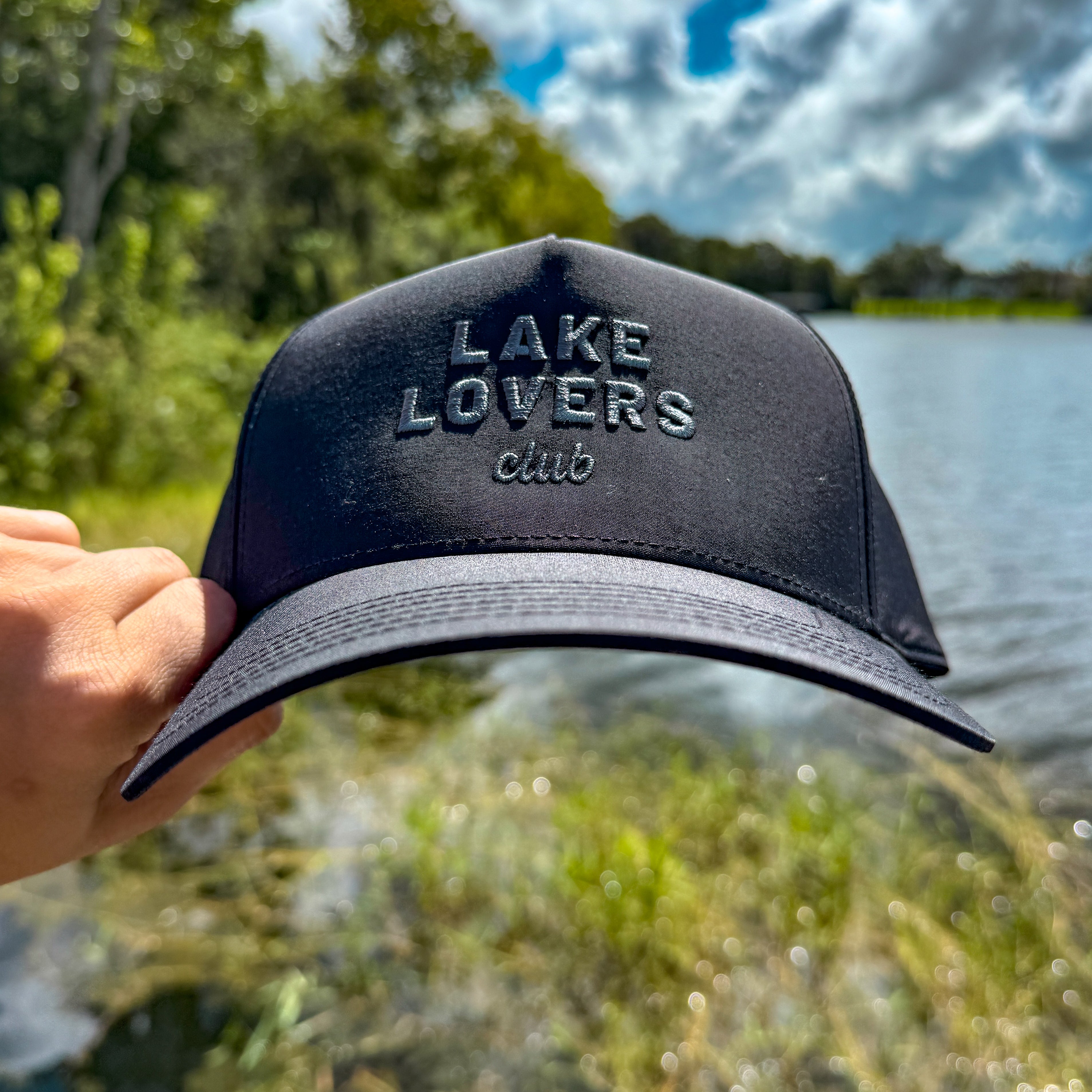 Black cap with 'Lake Lovers club' text held over a scenic lake and greenery background