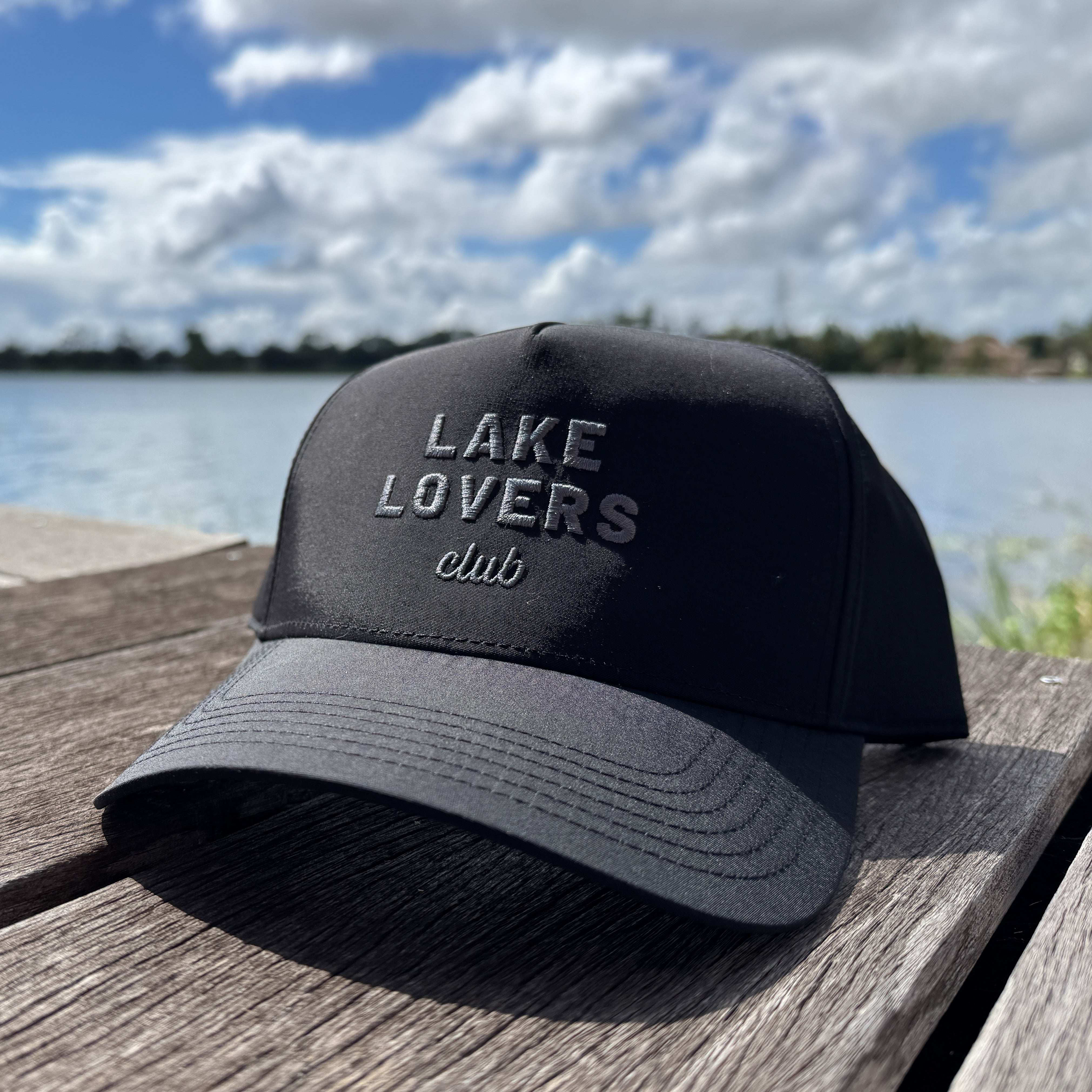 Black cap with 'Lake Lovers Club' text on a wooden surface by a lake.