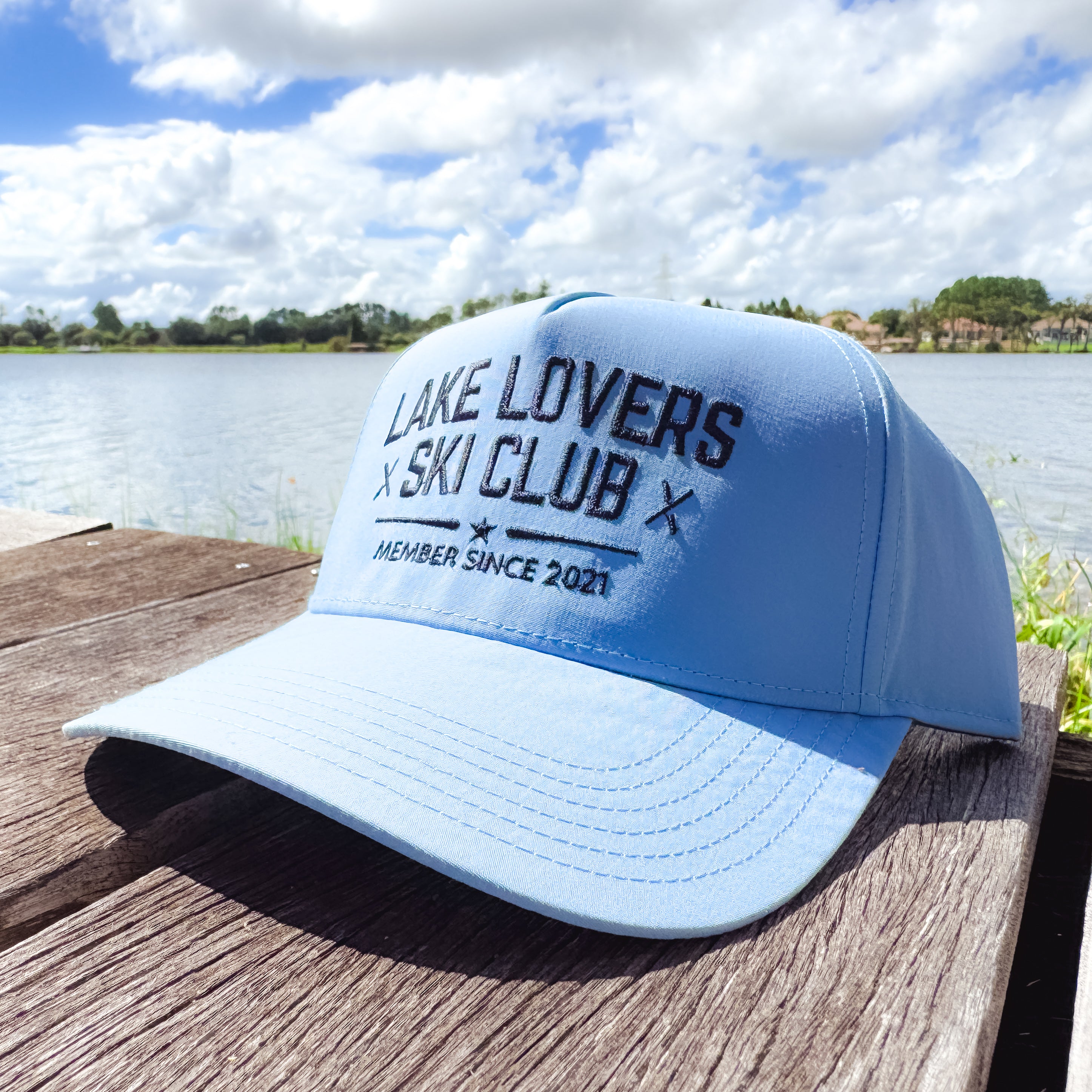Light blue cap with 'Lake Lovers Ski Club' text on a wooden dock by a lake.