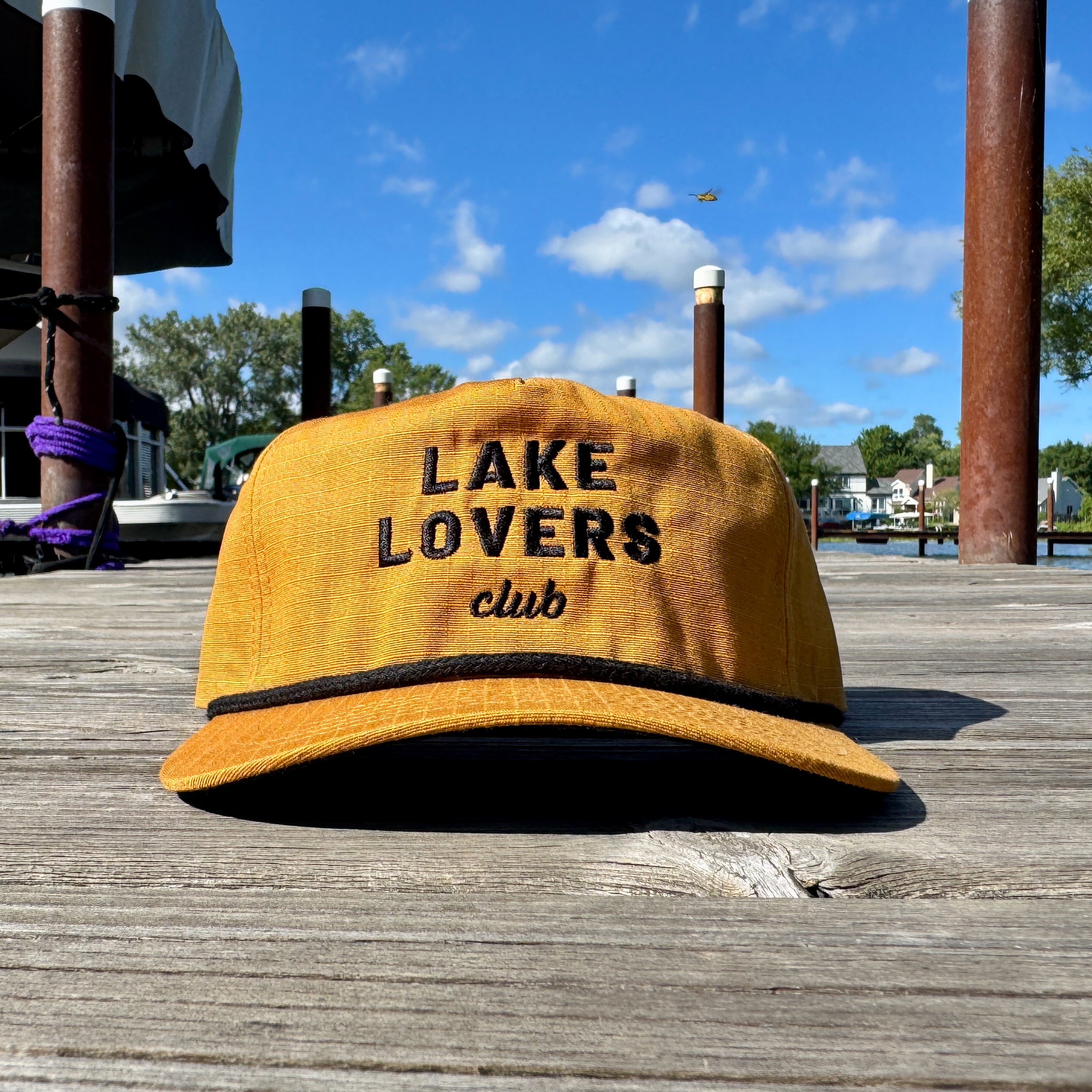 Yellow cap with 'Lake Lovers Club' text on a wooden dock with a lake and trees in the background.
