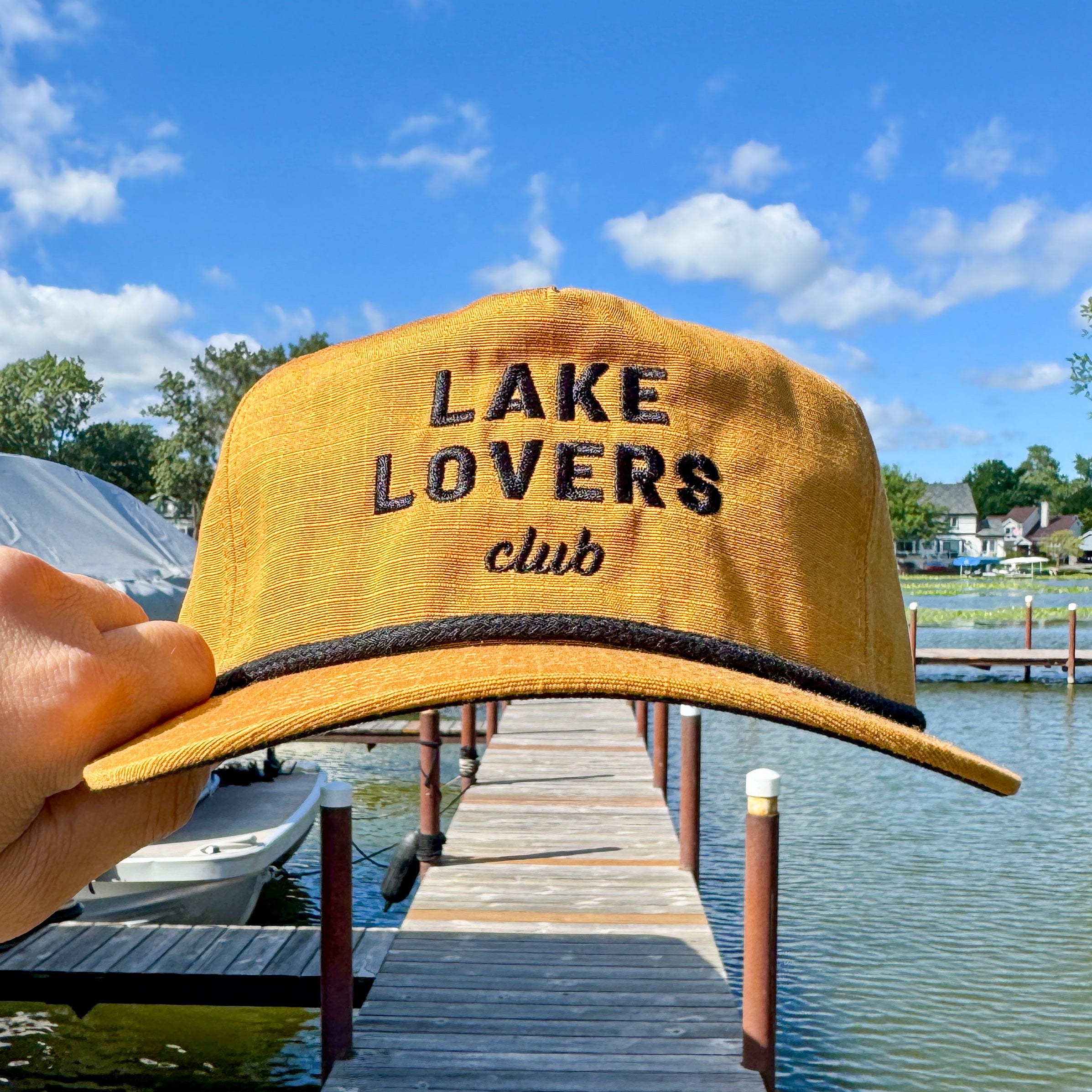 Yellow cap with 'Lake Lovers Club' text held over a lake with docks and boats.