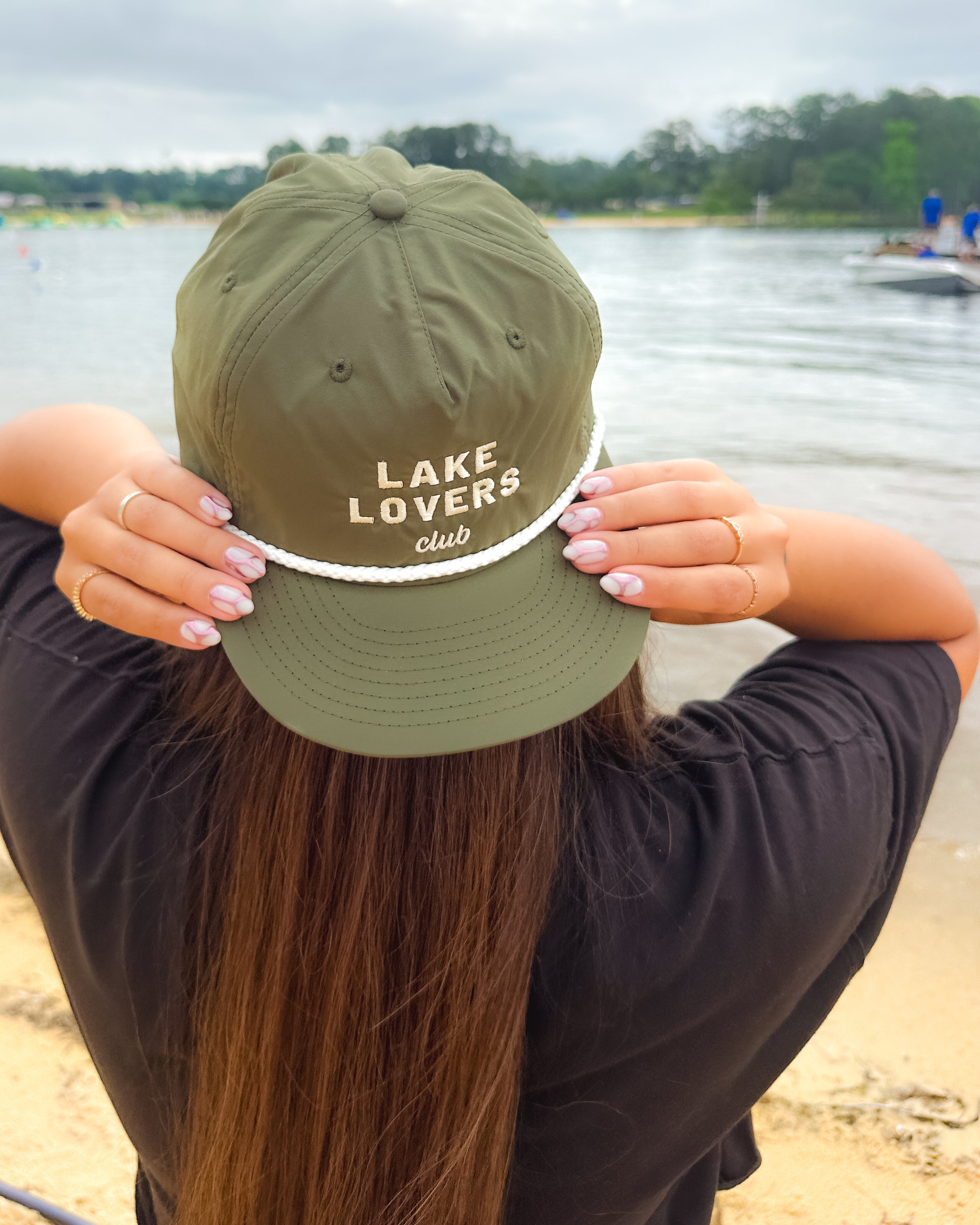 Woman wearing a backwards olive green cap with 'Lake Lovers Club' text by a lake.