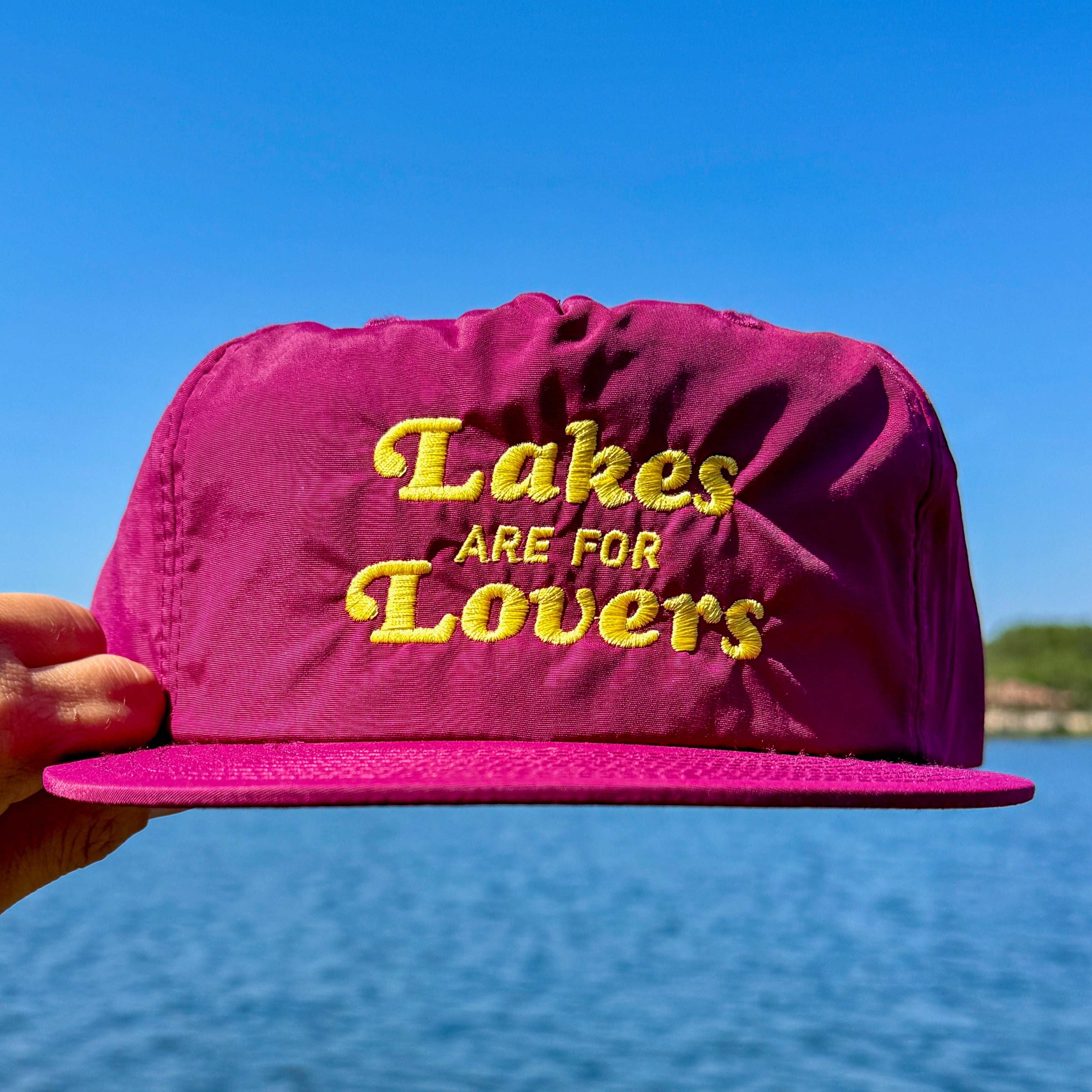 Maroon cap with 'Lakes are for Lovers' text held over a lake