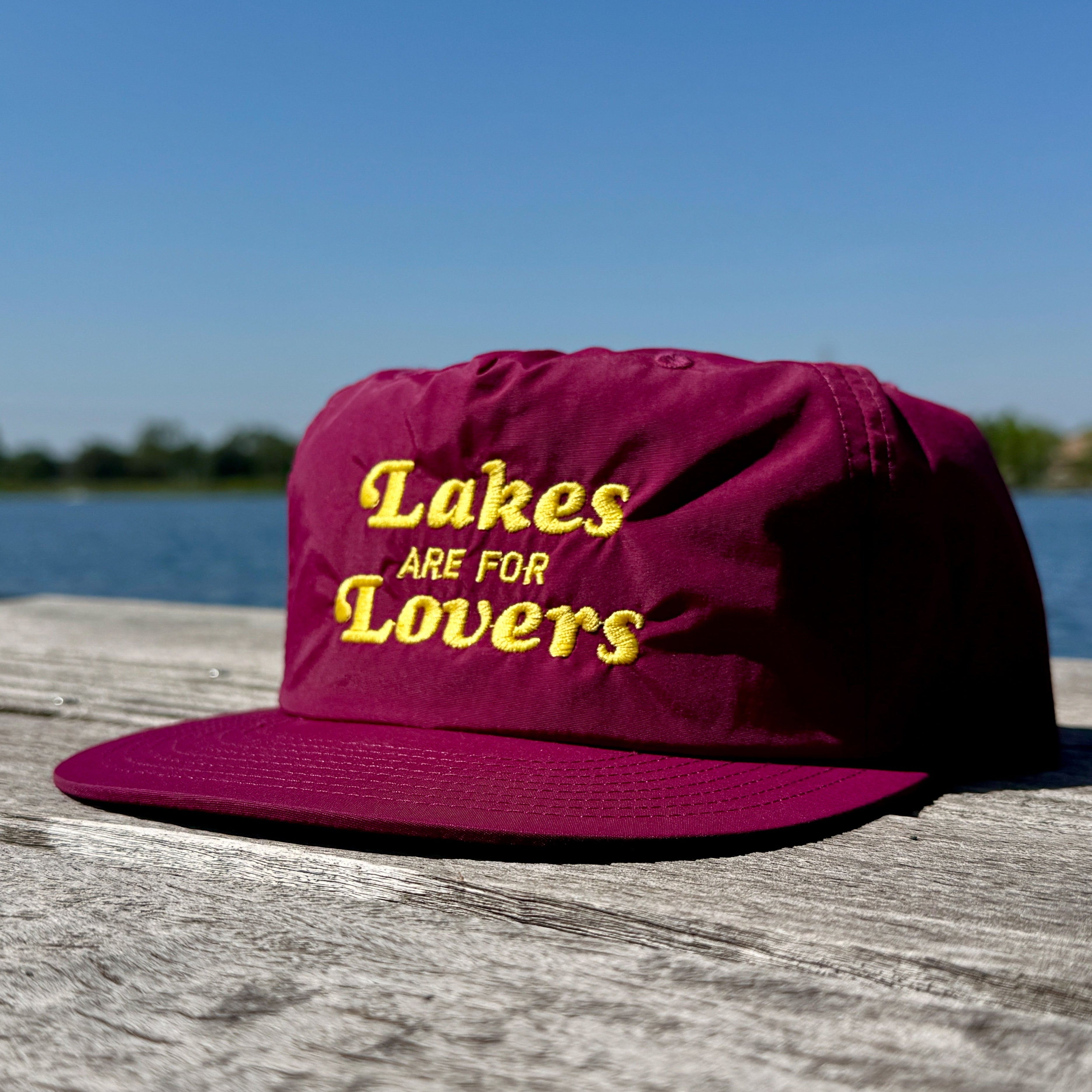 Maroon cap with 'Lakes are for Lovers' text on a wooden surface with a lake background