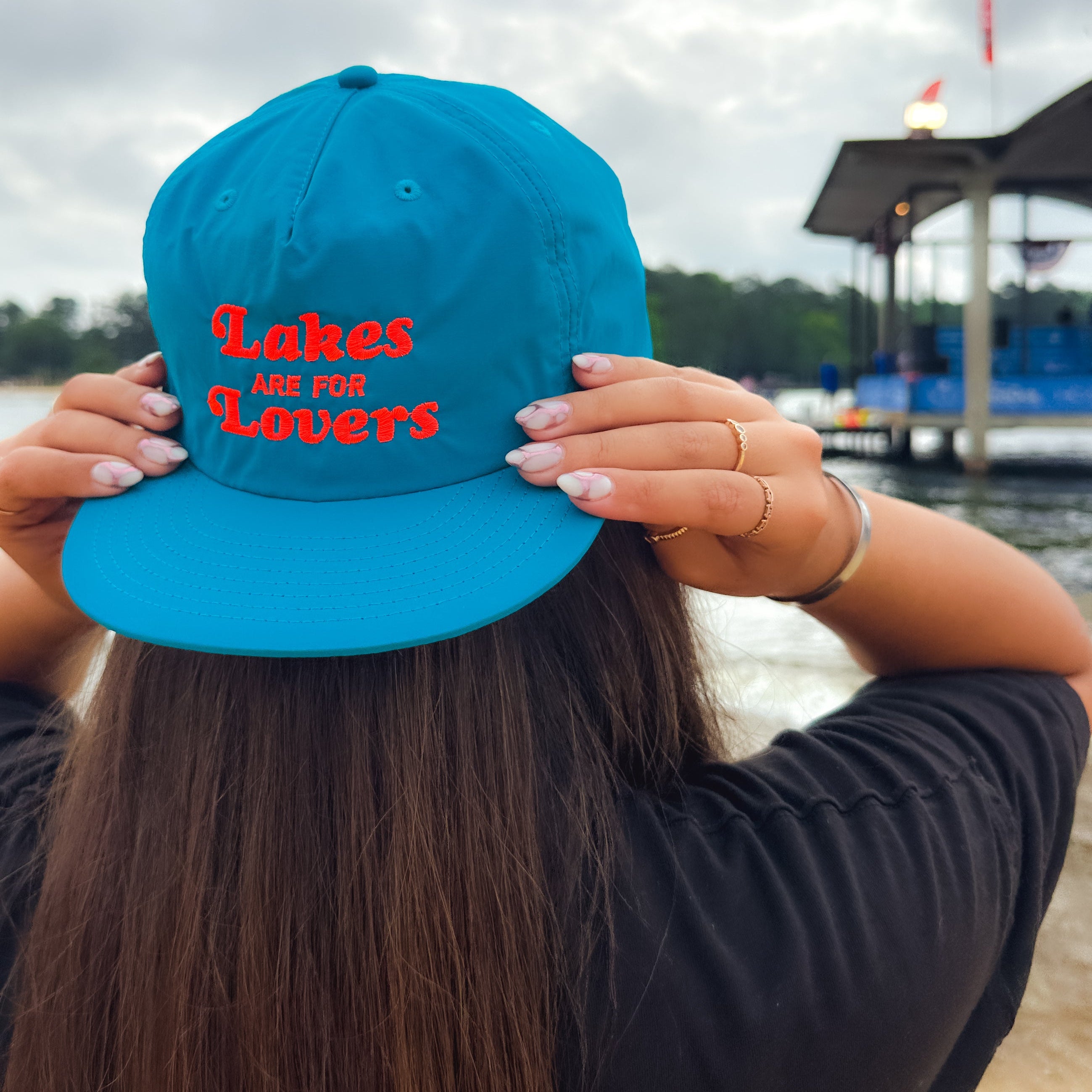 Person wearing a blue cap with 'Lakes are for Lovers' text by a lake.