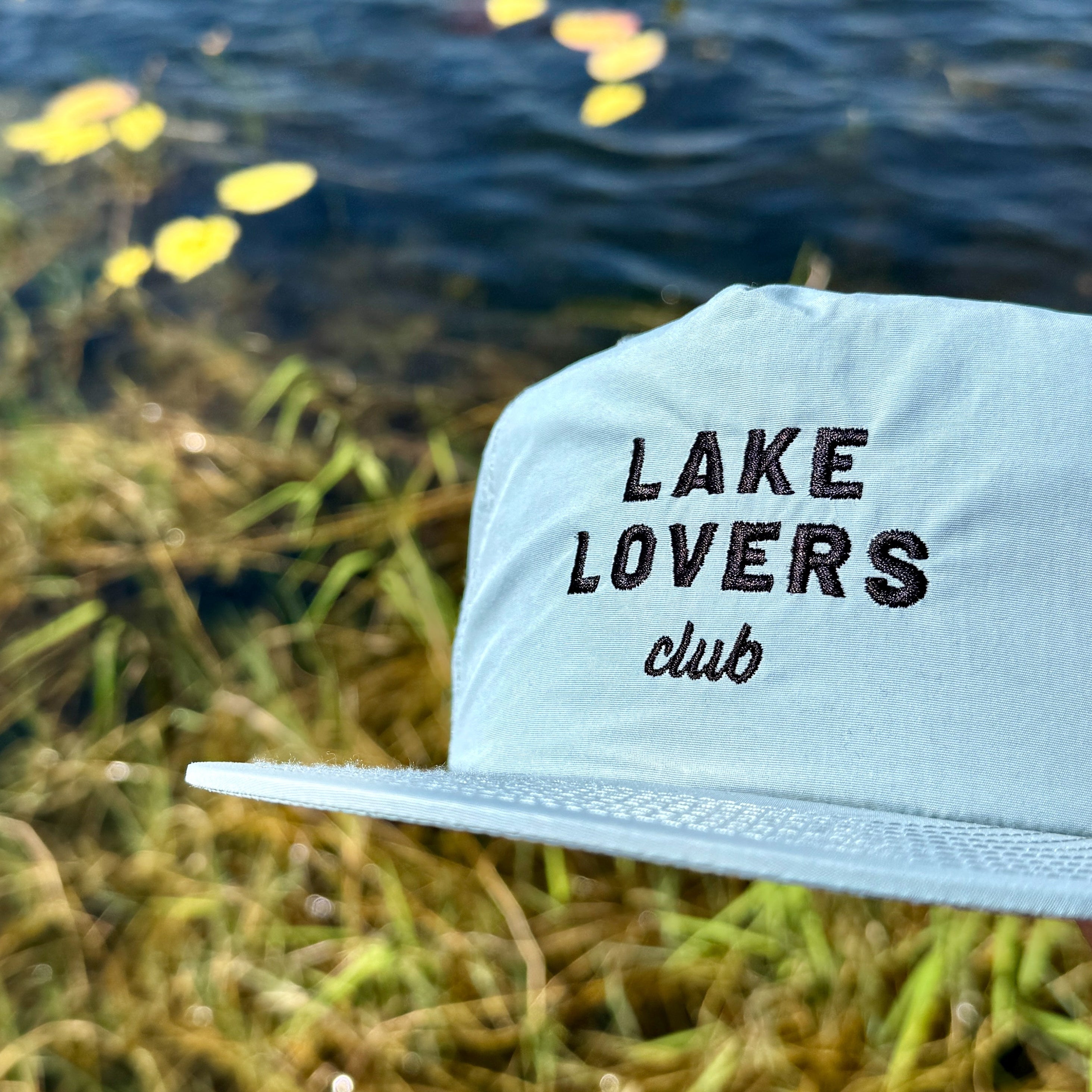 Light grey cap with 'Lake Lovers Club' text against a natural background with water and plants.