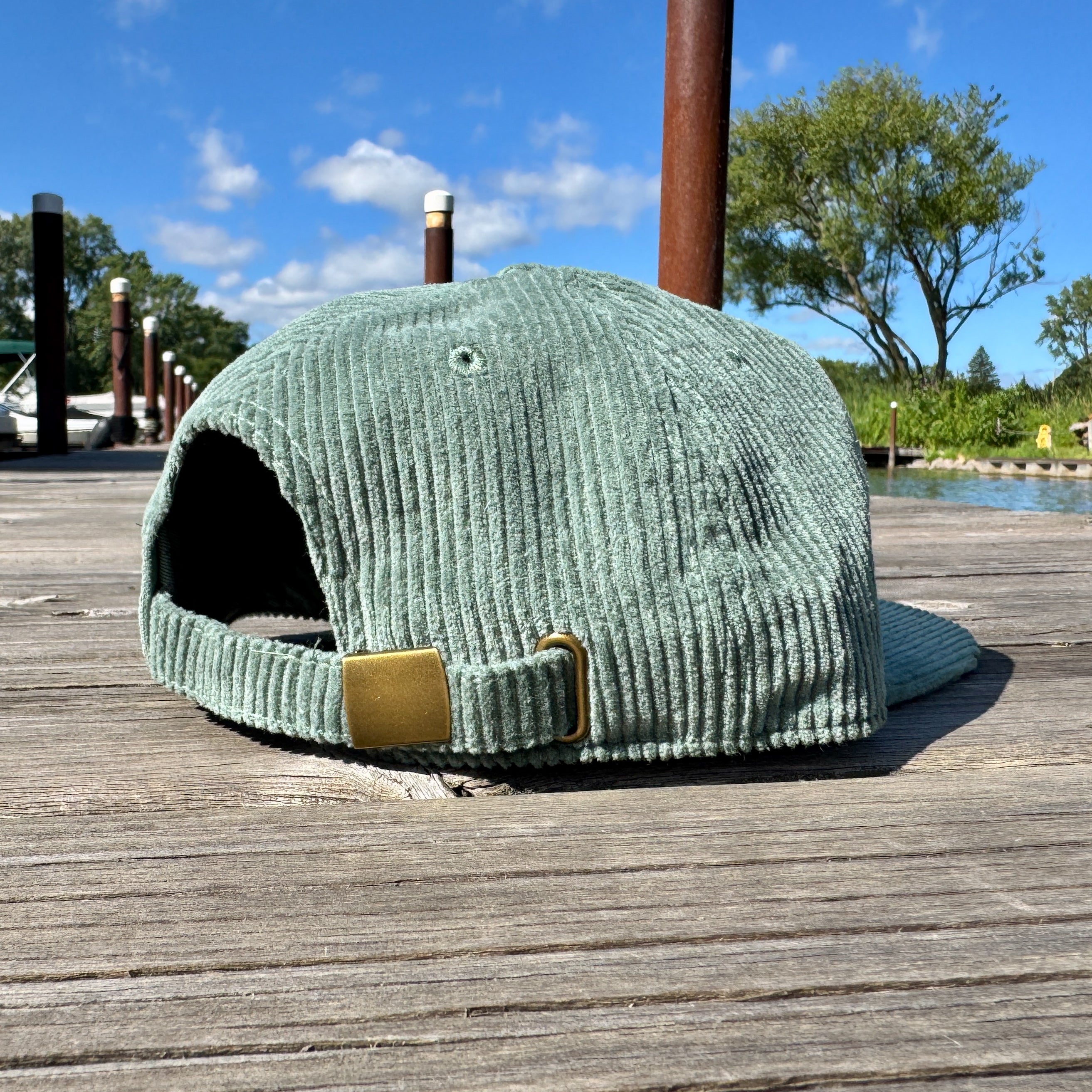 Green corduroy cap with a gold buckle on a wooden dock with a scenic background.