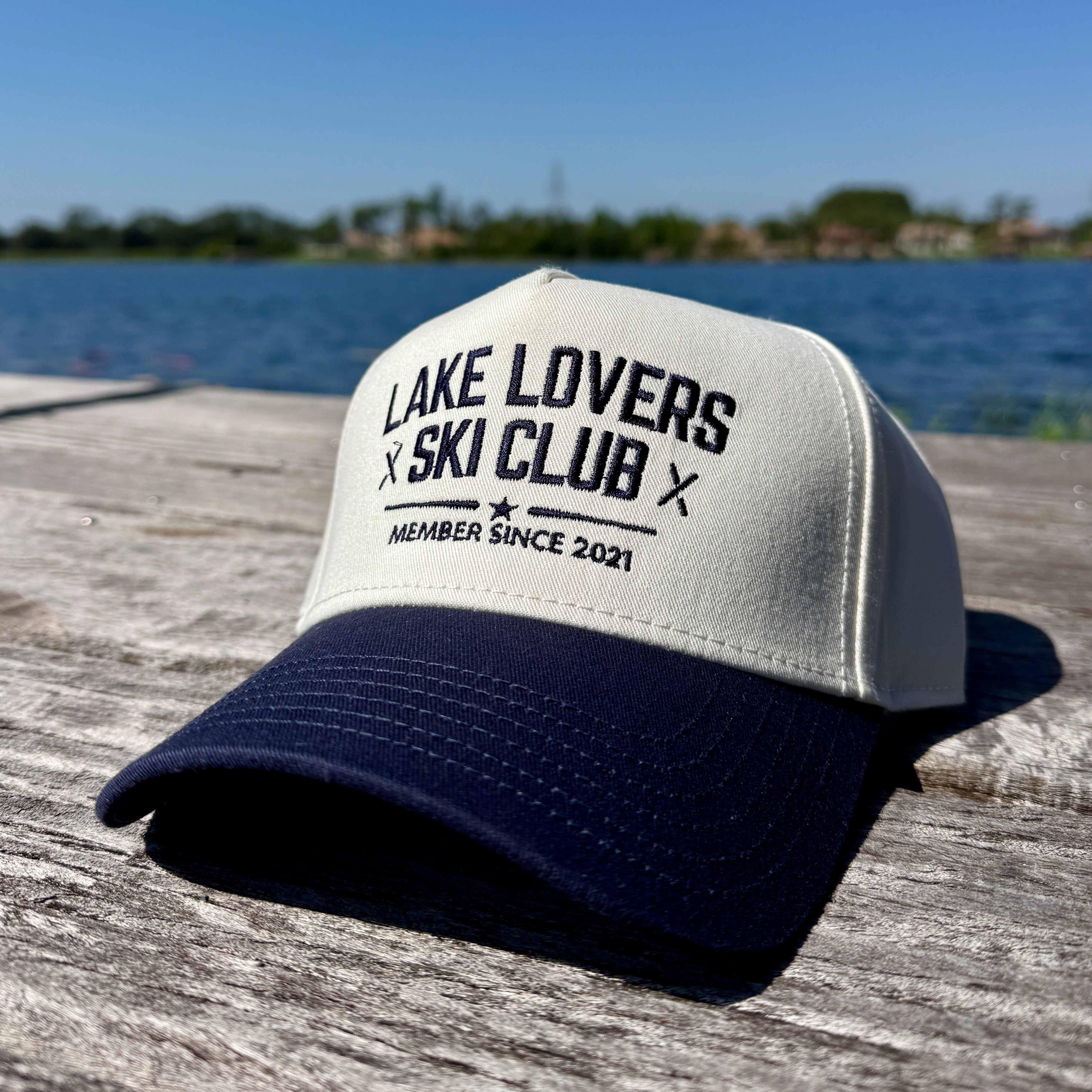 Two-tone baseball hat with 'Lake Lovers Ski Club' text on a wooden dock with a lake background.