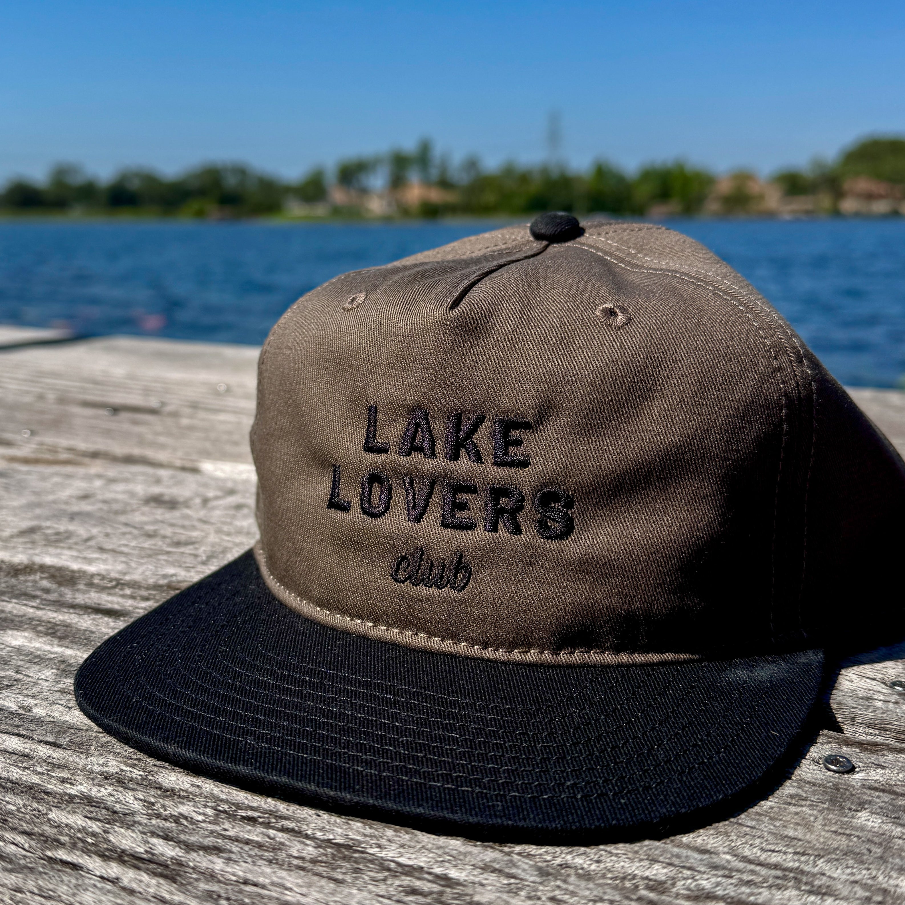 Brown and black cap with 'Lake Lovers Club' text on a wooden dock with water and sky in the background