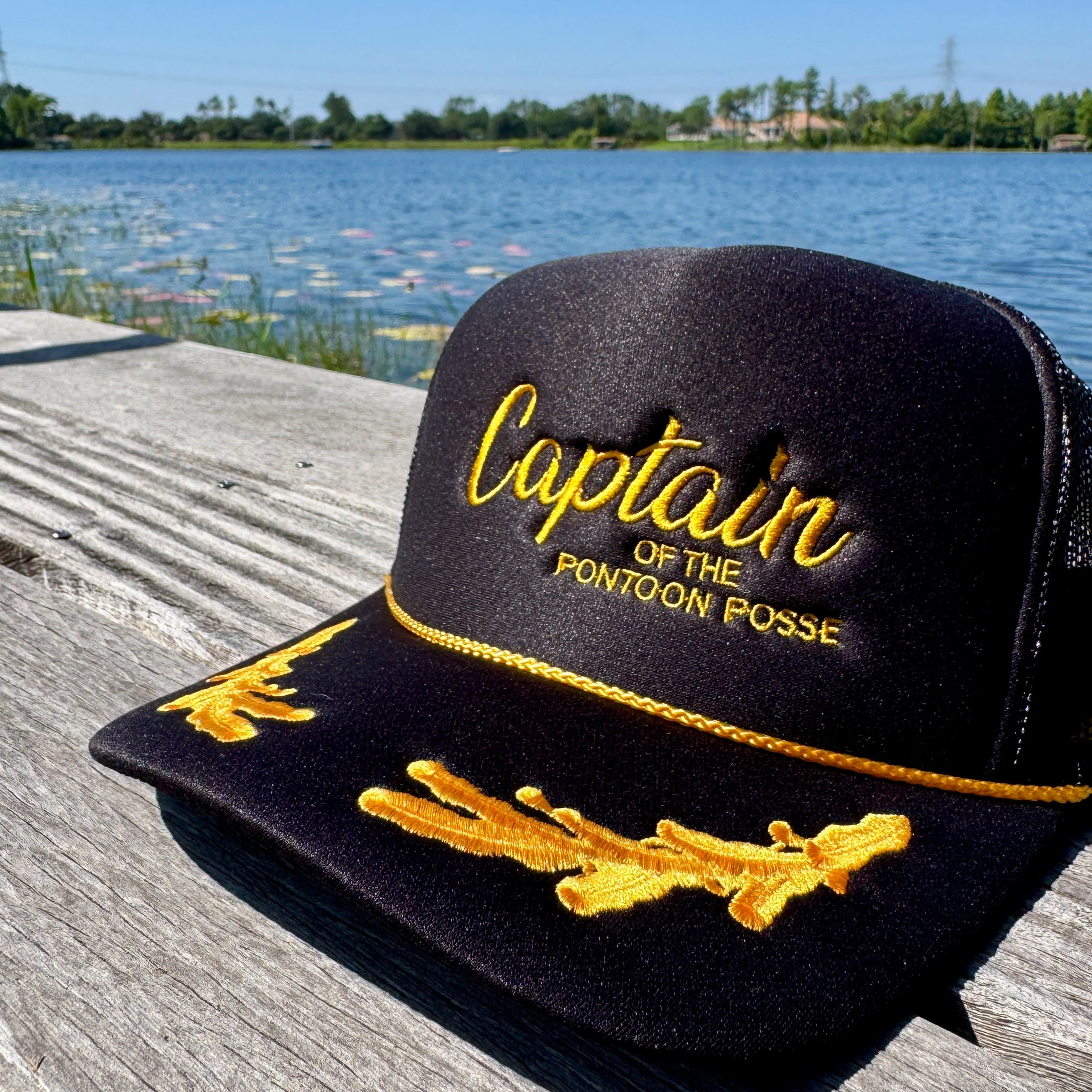 Black cap with 'Captain of the Pontoon Posse' text on a wooden dock by a lake.