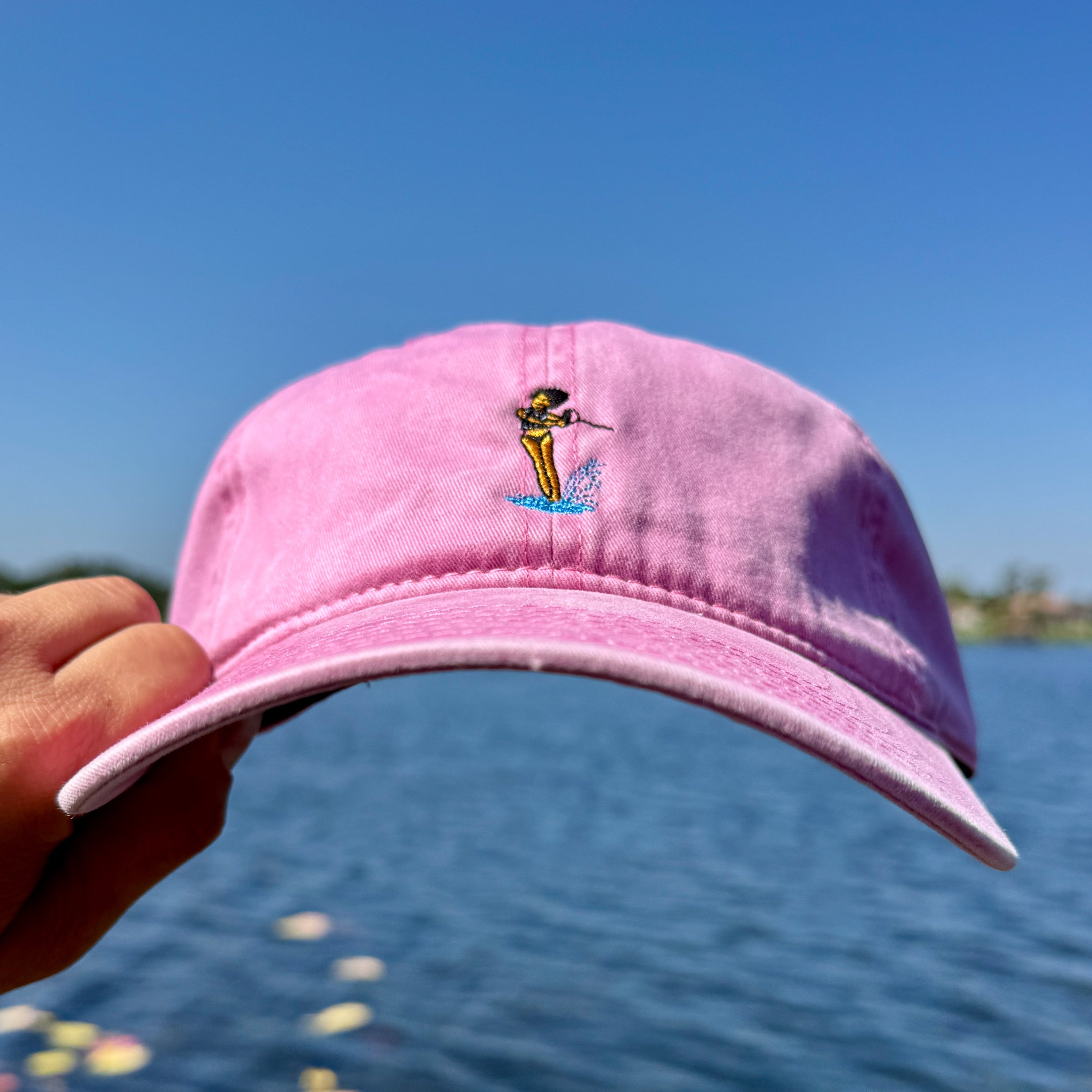 Hand holding a pink hat in front of a lake, with a waterski girl embroidered on the front of it.