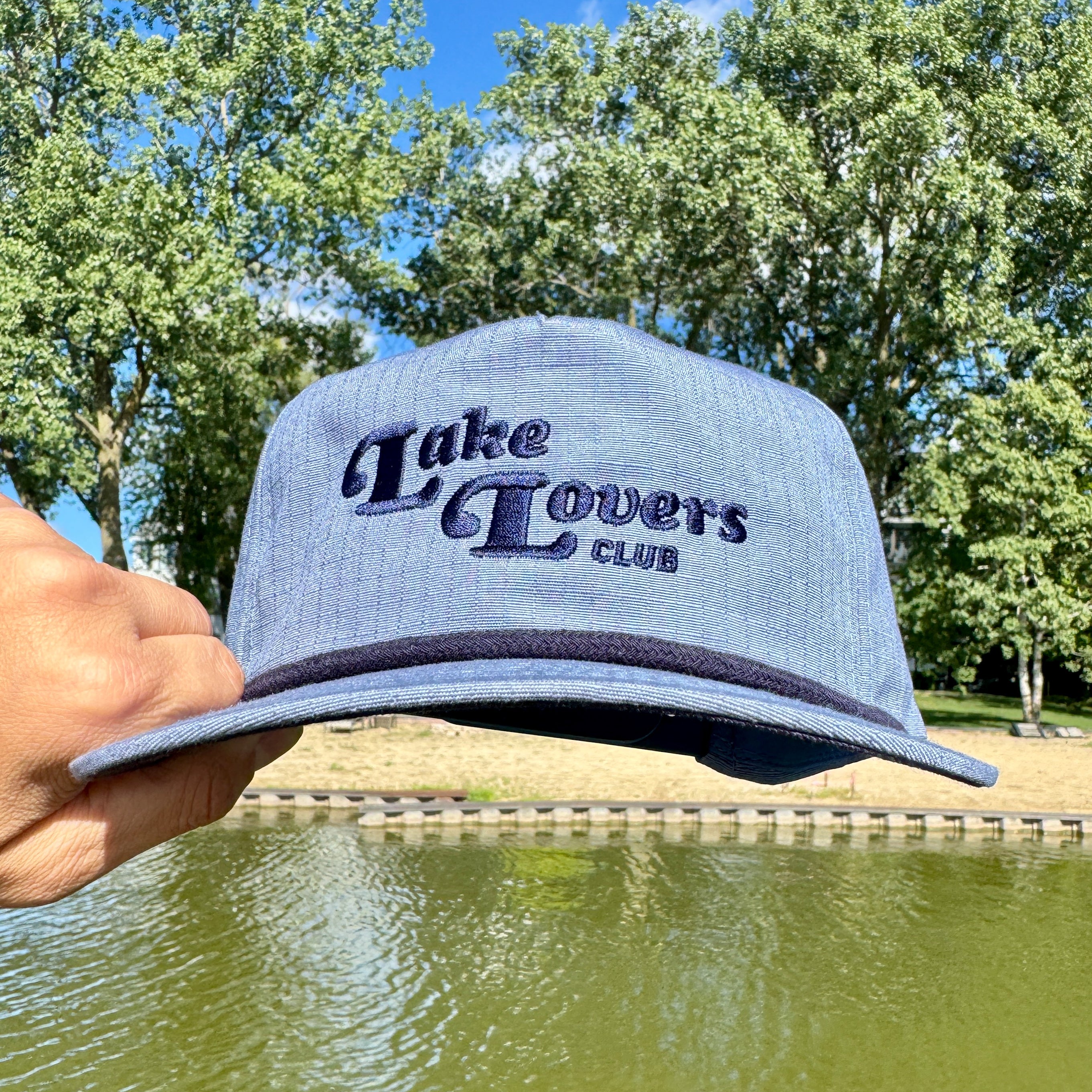 Blue cap with 'Lake Lovers Club' text held by a hand with a lake and trees in the background