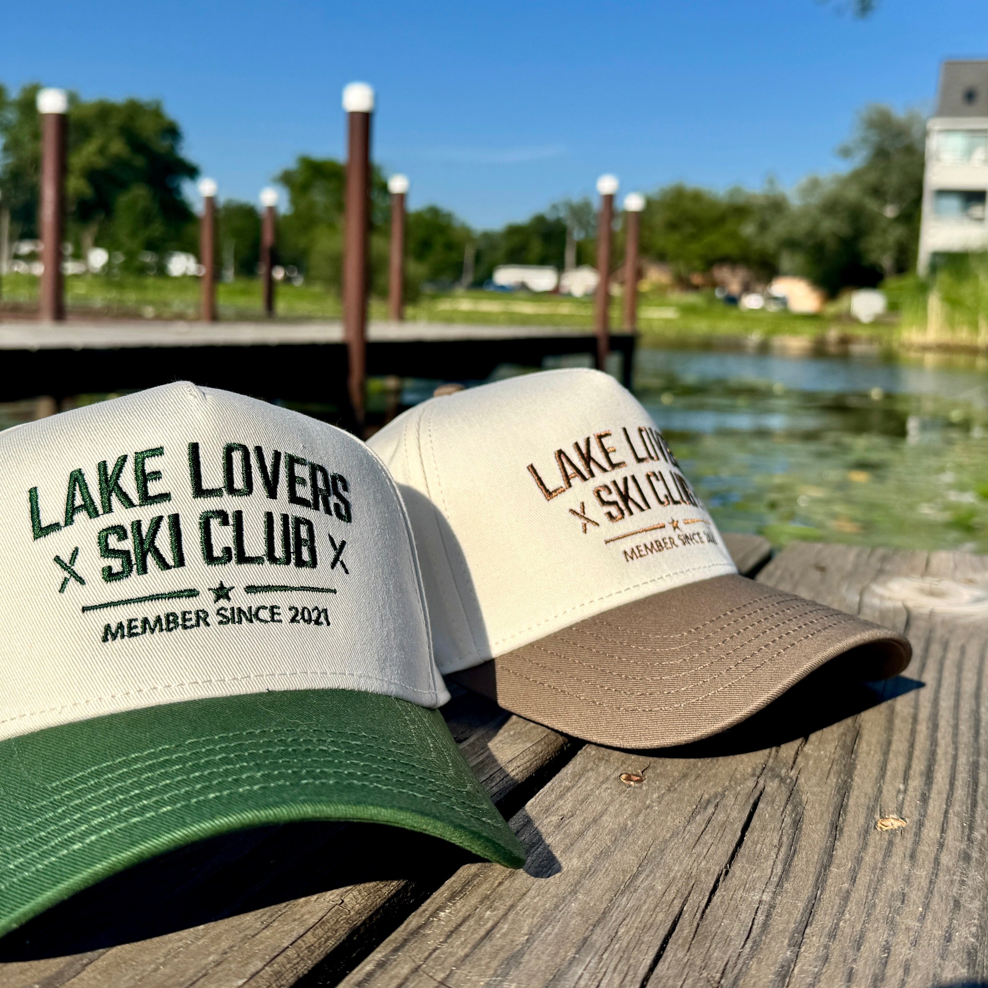 Two beige hats on a dock. One has a forest green brim and the other has a brown. Both have text on them that says Lake Lovers Ski Club.