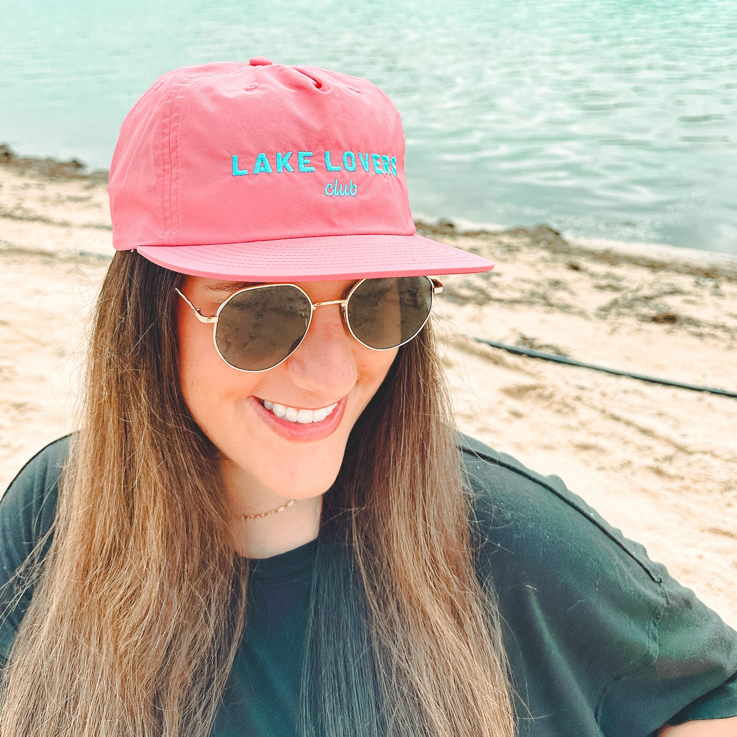 Woman wearing a pink 'Lake Lover' cap and sunglasses by a lake.