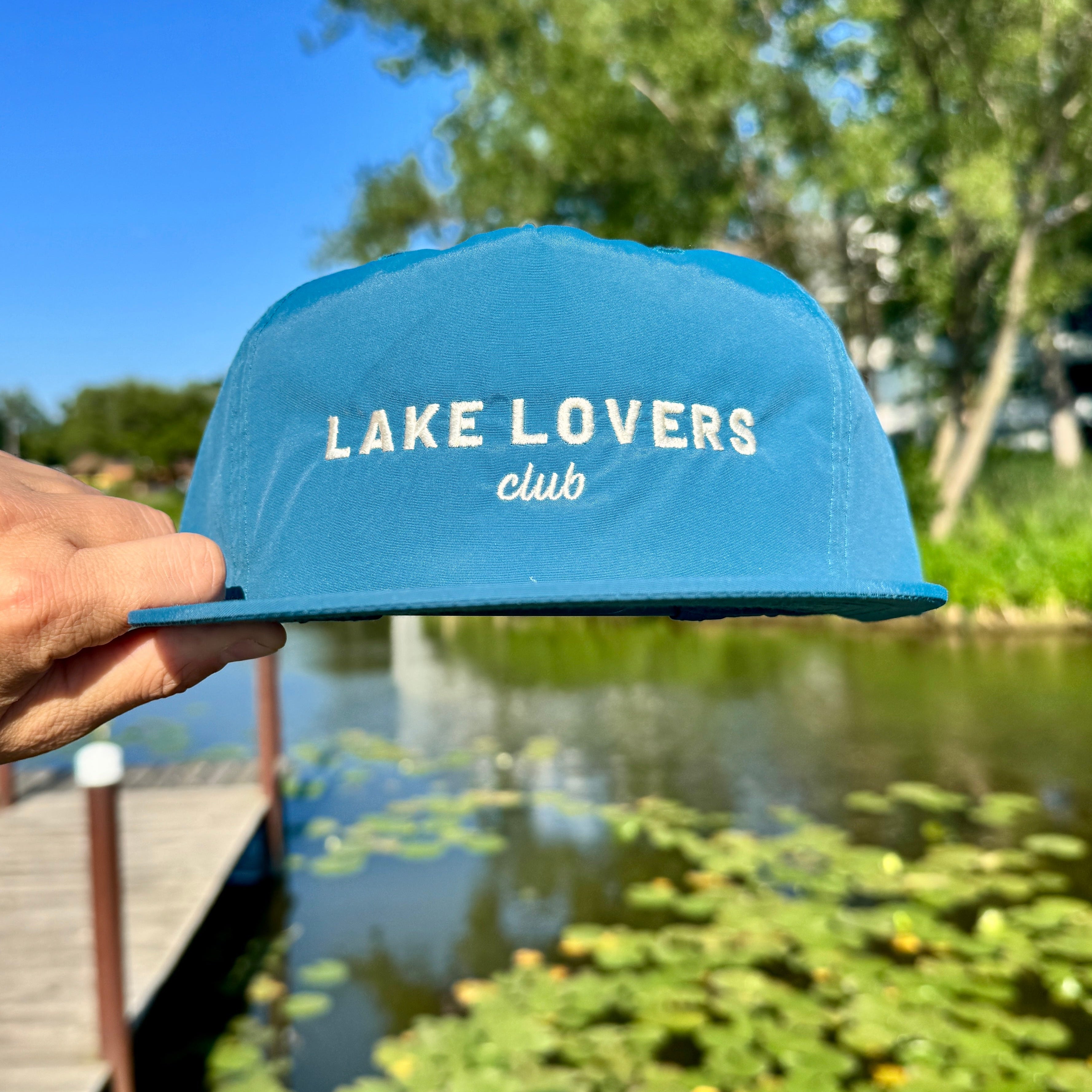 Hand holding a teal quick dry nylon hat over water, with the Lake Lovers Club logo on the front of the hat.