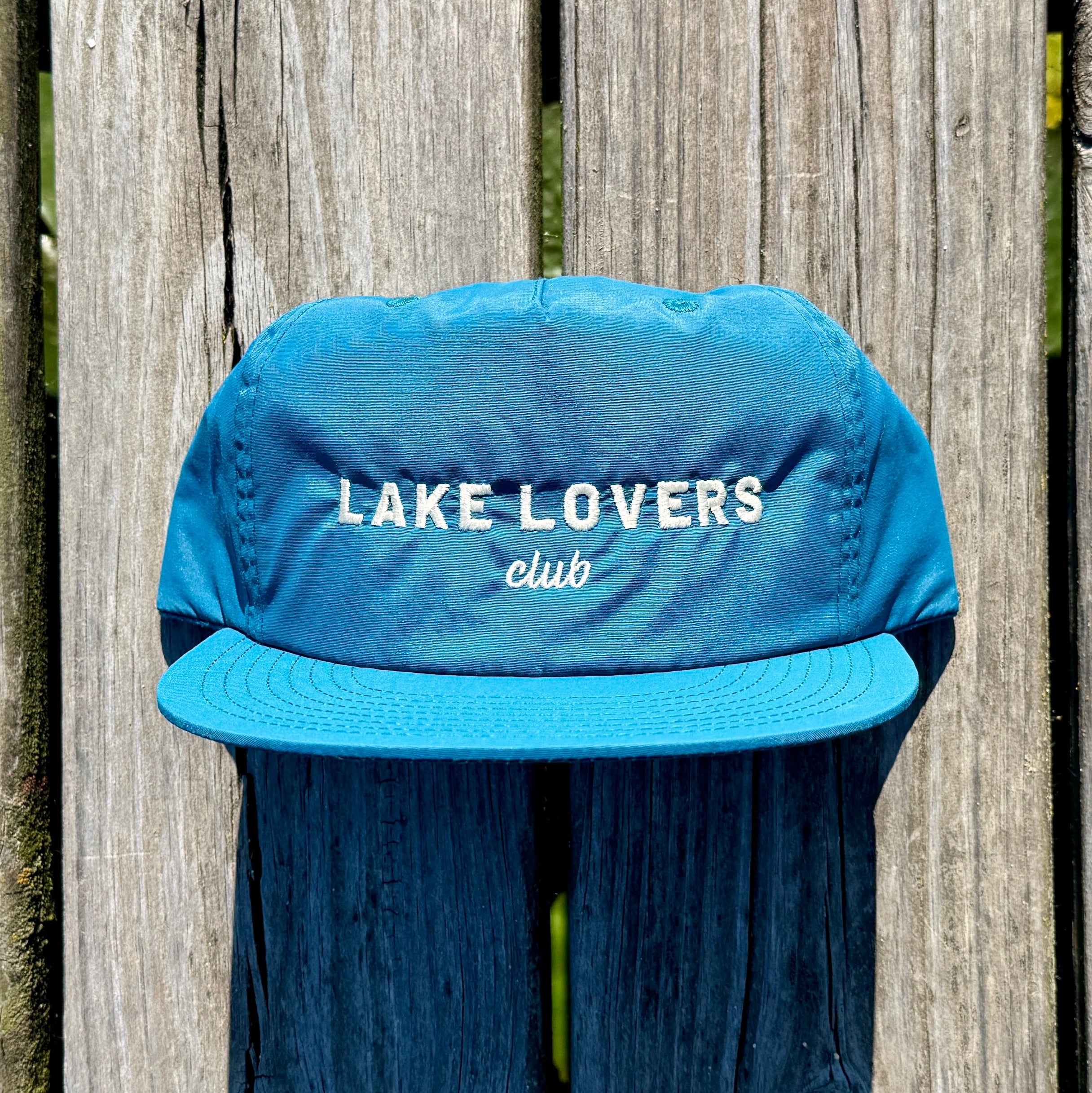 A teal quick dry nylon rope hat sitting on a dock. That hat says Lake Lovers Club in white thread.