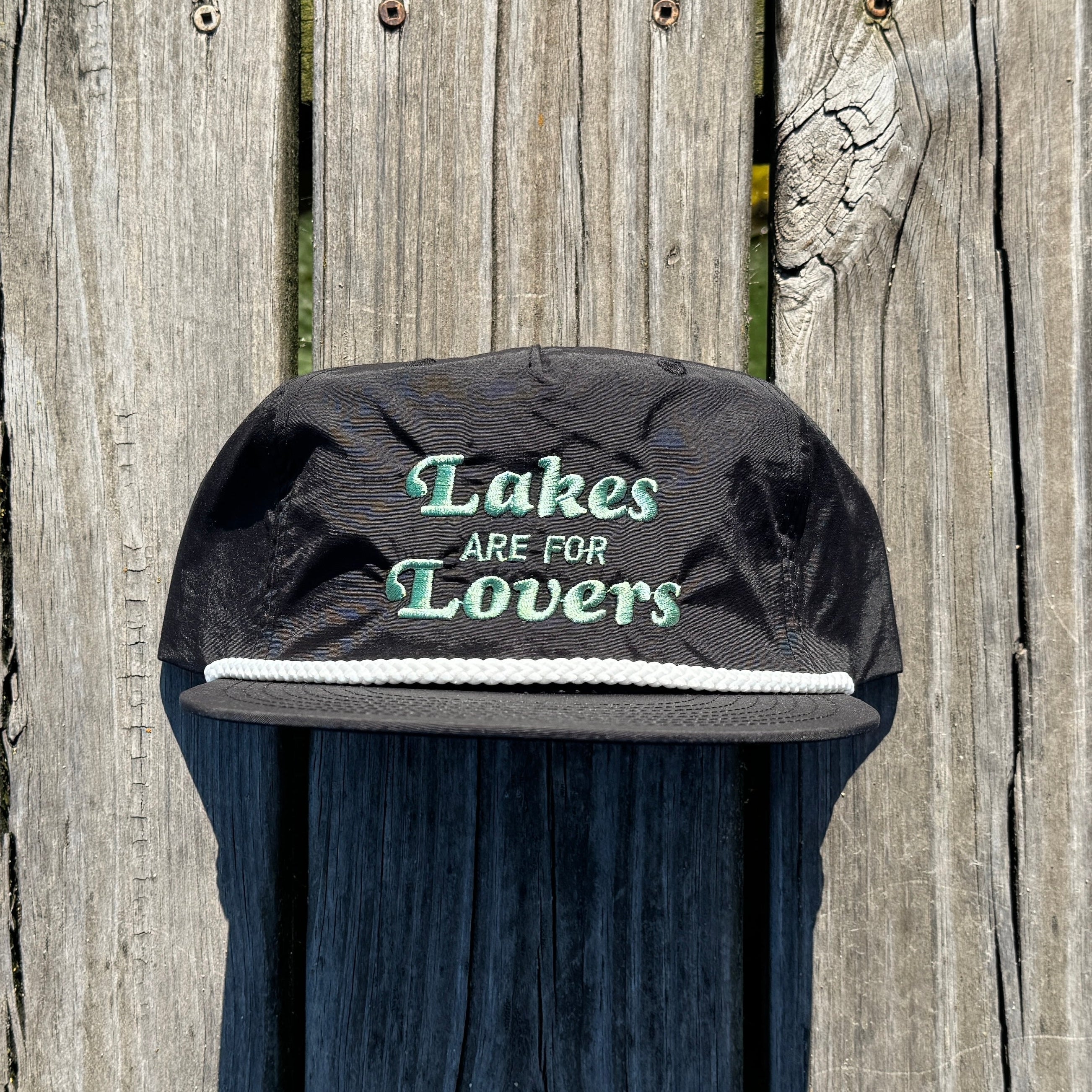 A black quick dry nylon rope hat sitting on a dock. That hat says Lakes are for Lovers in olive green thread.
