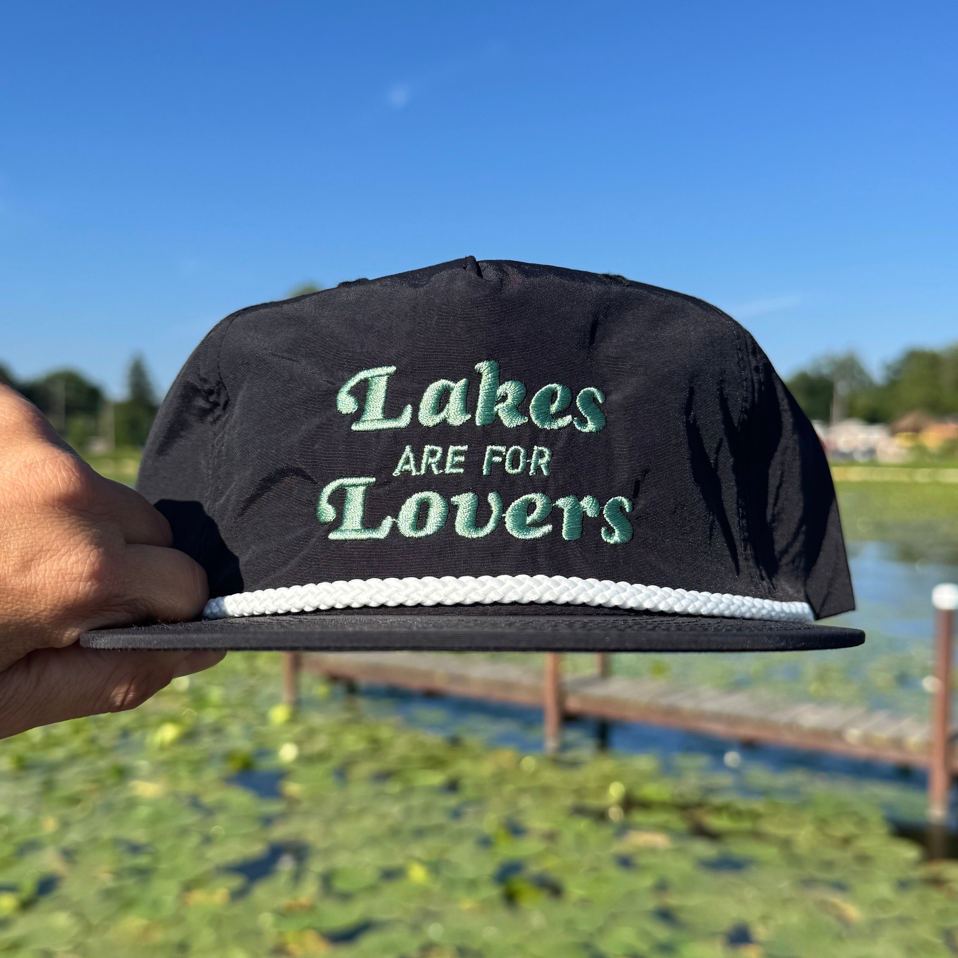 Hand holding a black quick dry nylon rope hat over water that says Lakes are for Lovers in blue thread.