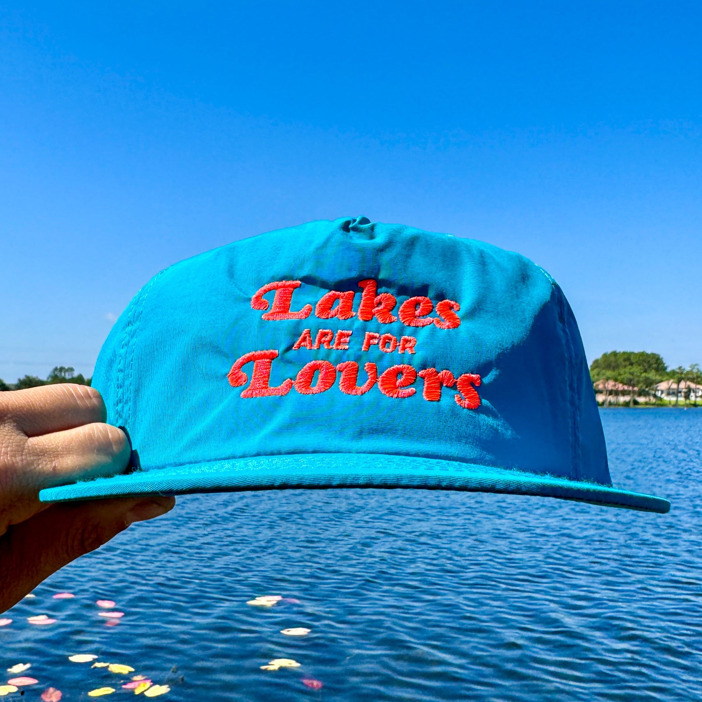 teal hat that reads 'lakes are for lovers,' held by a hand in front of a body of waer