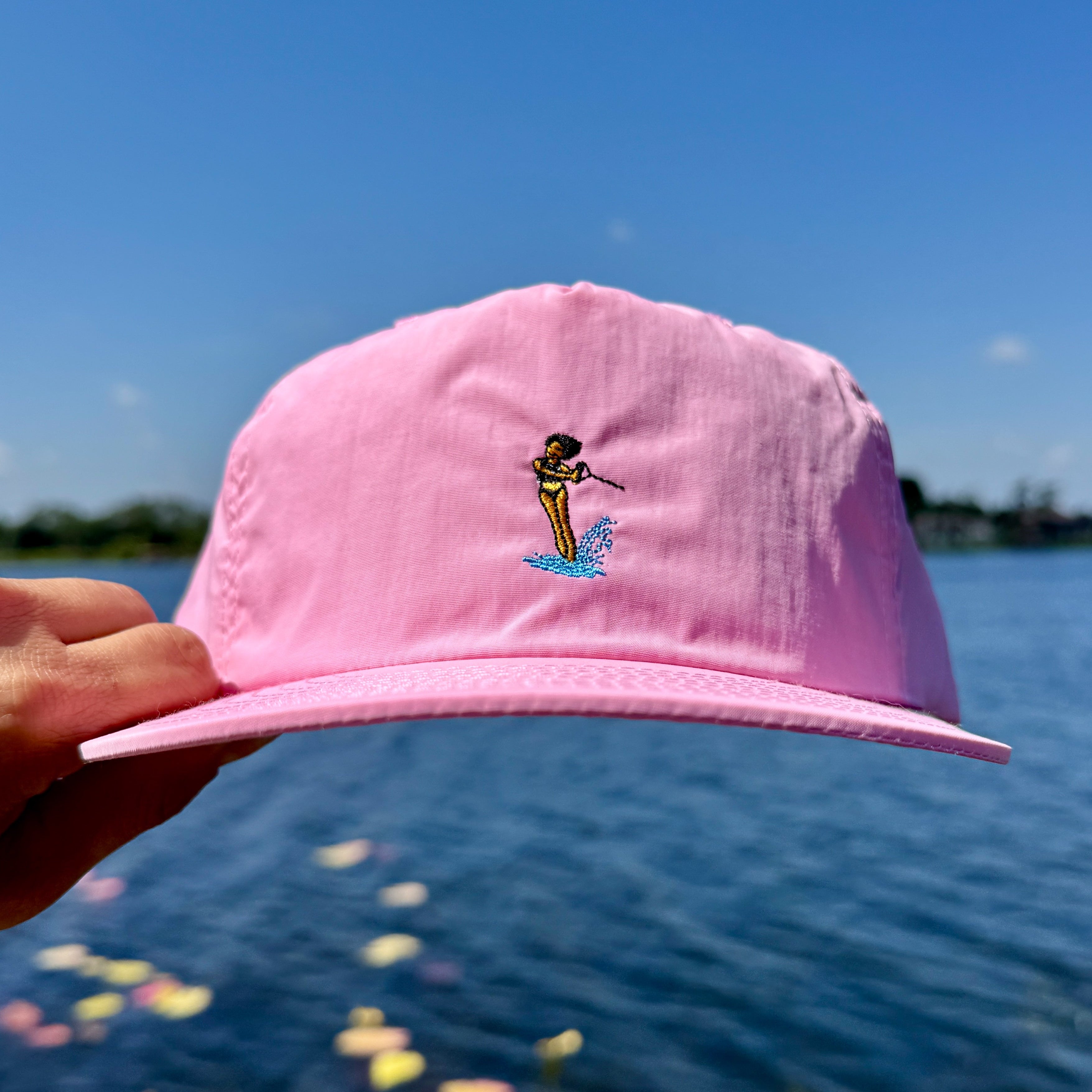 Pink cap with a waterski gril design held up against a blue sky and water background