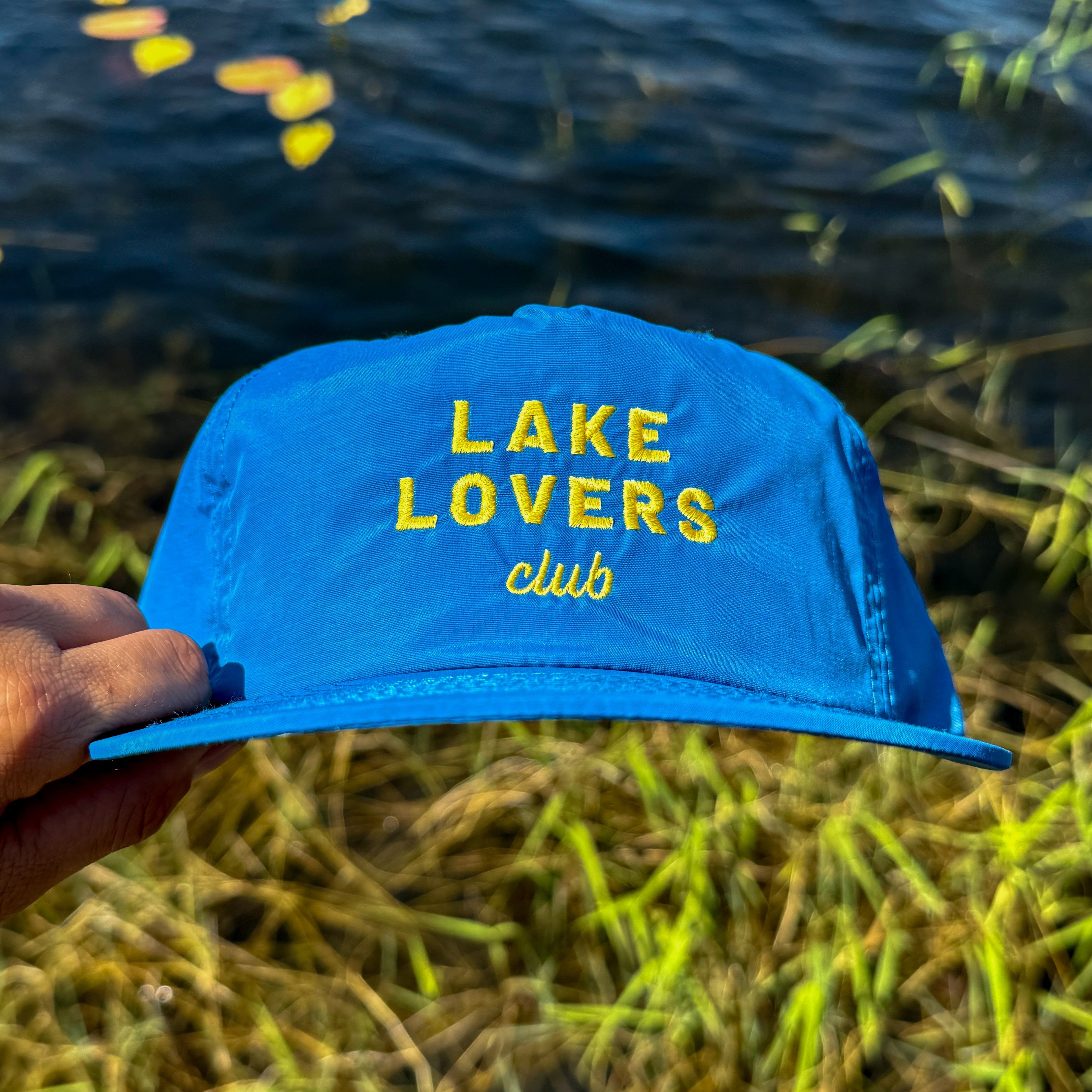 Blue cap with 'Lake Lovers club' text held over a lake with water lilies.