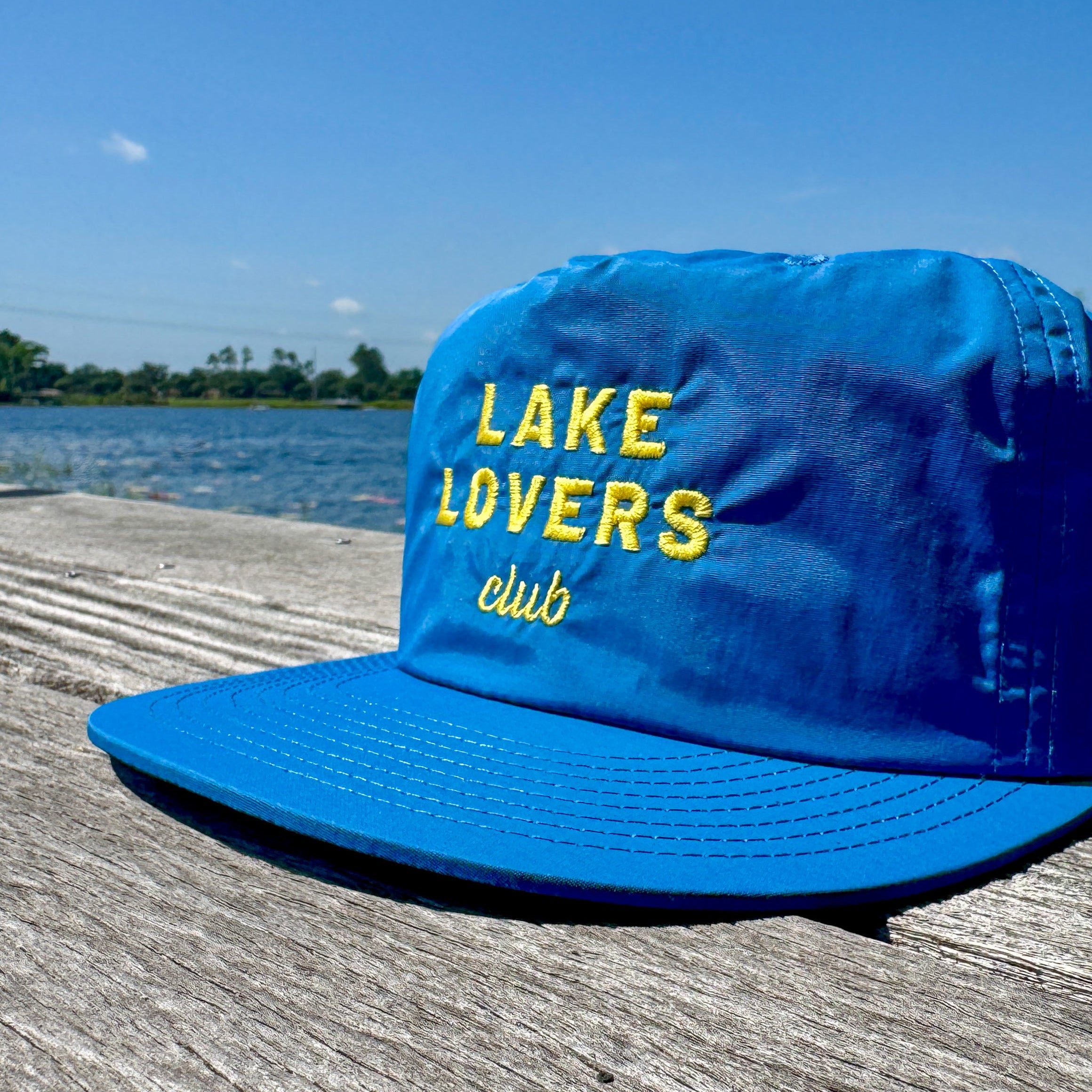 Blue cap with 'Lake Lovers club' text on a wooden dock by a lake.
