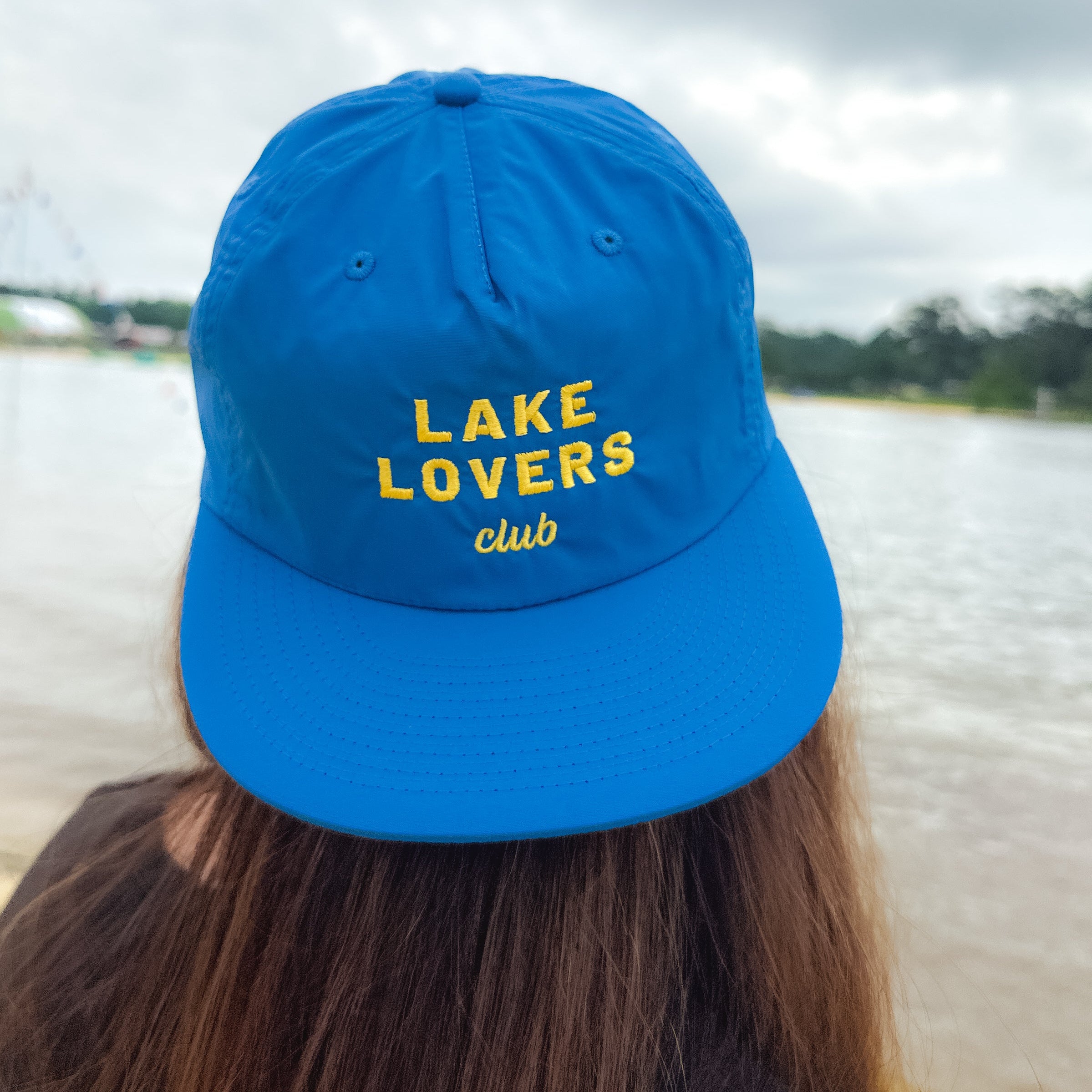 Person wearing a blue cap with 'Lake Lovers Club' text by a lake.
