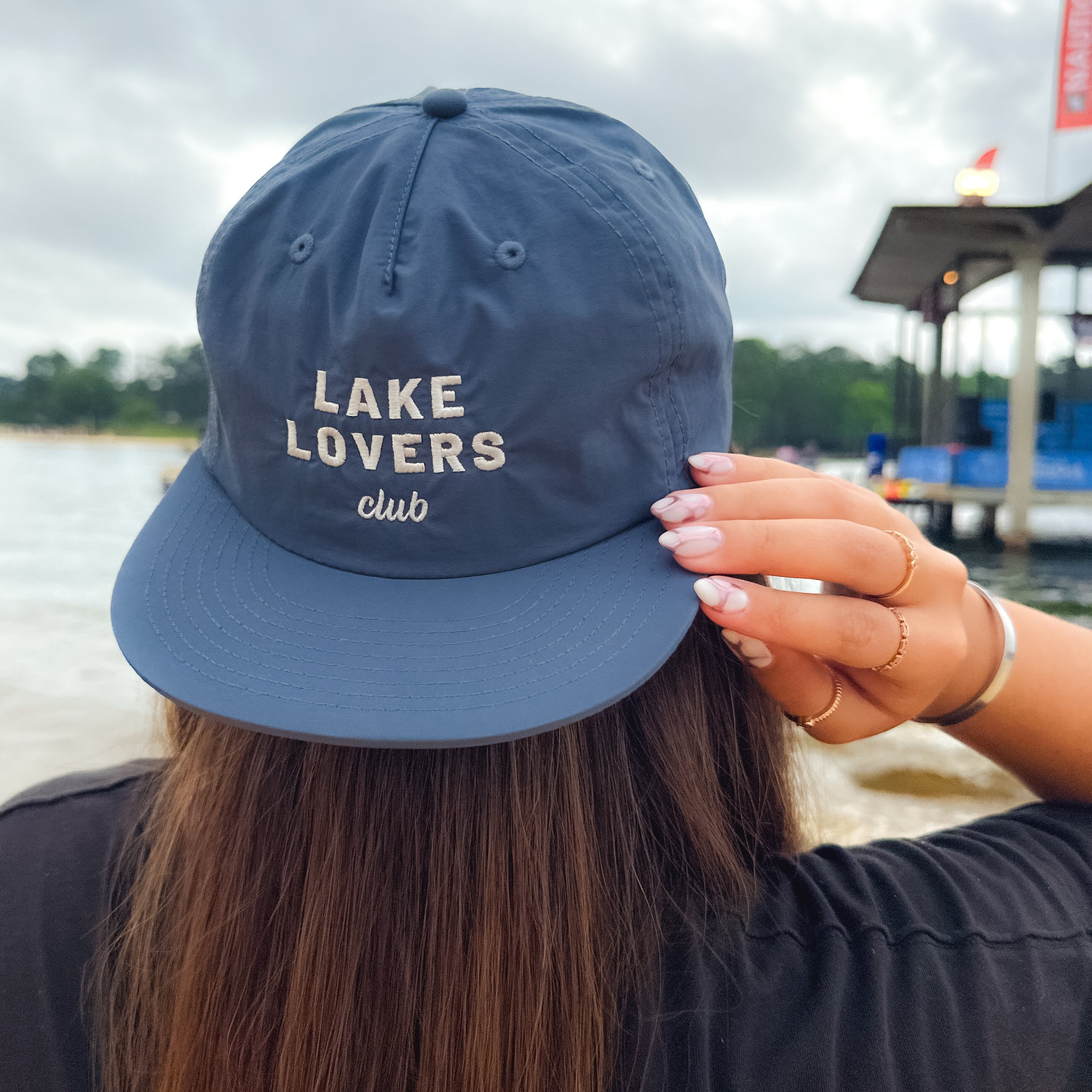 Person wearing a navy blue cap with 'Lake Lovers Club' text by a lake.