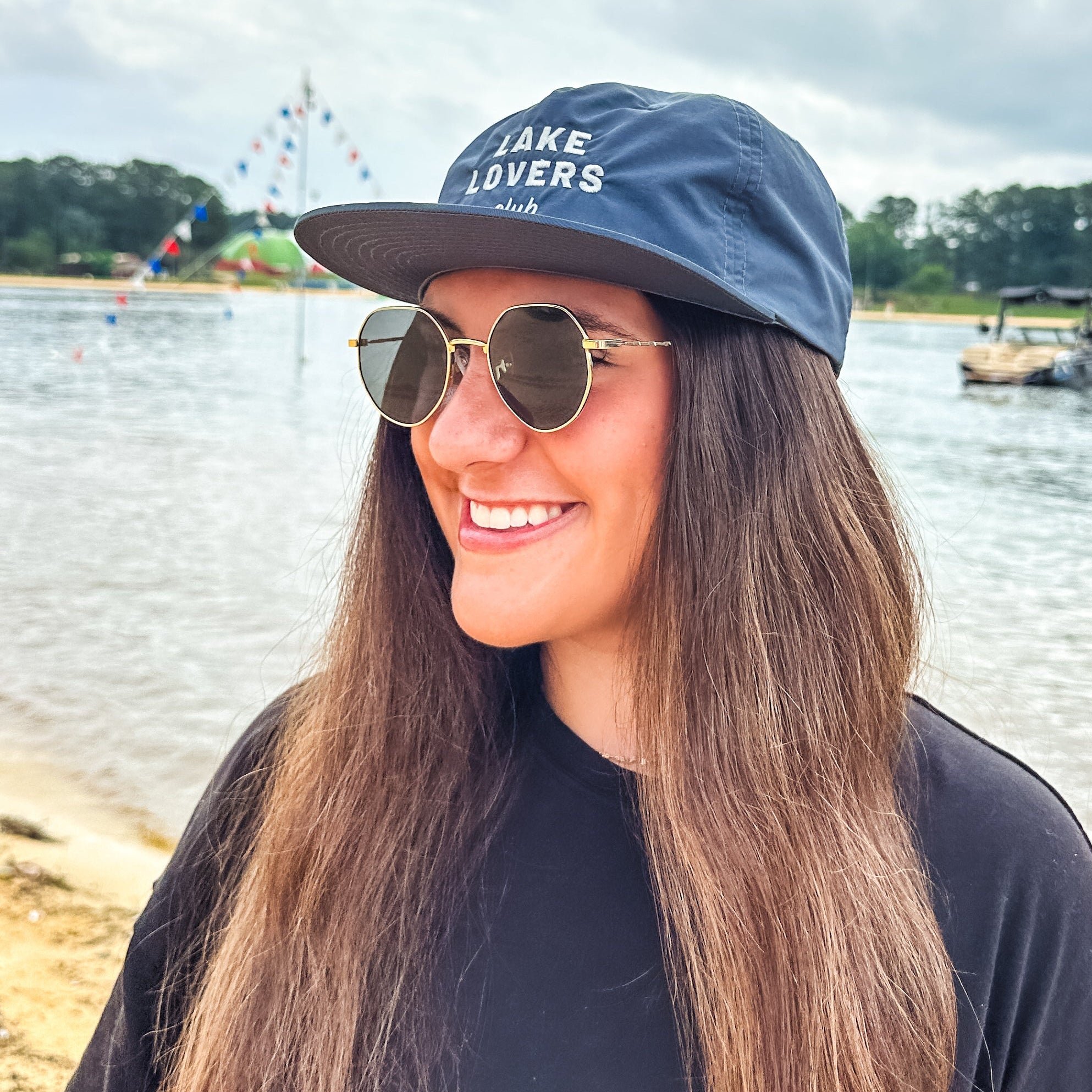 Woman wearing a 'Lake Lovers' cap and sunglasses by a lake with a pennant flags in the background.