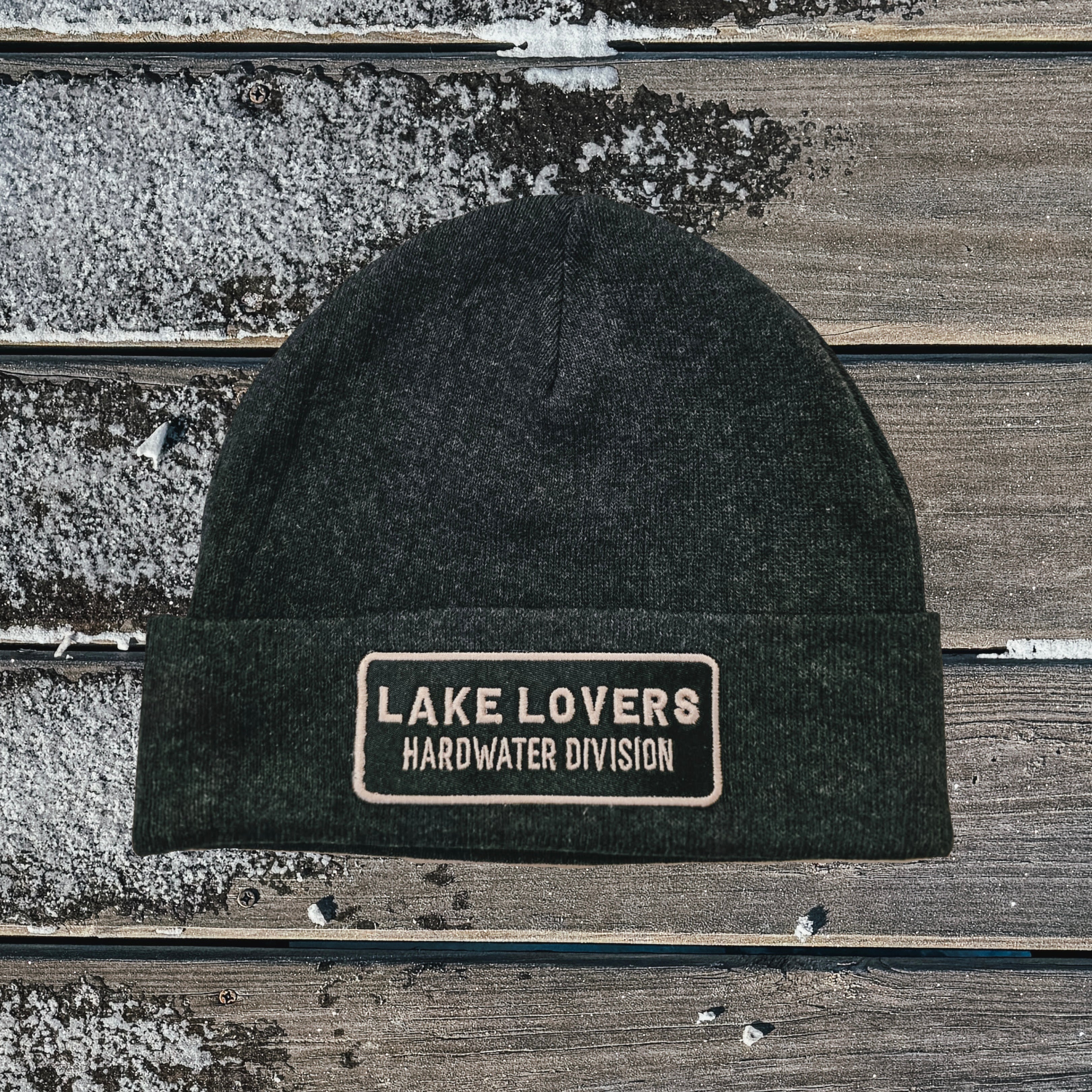 Dark gray beanie with 'Lake Lovers Hardwater Division' patch on a wooden surface.