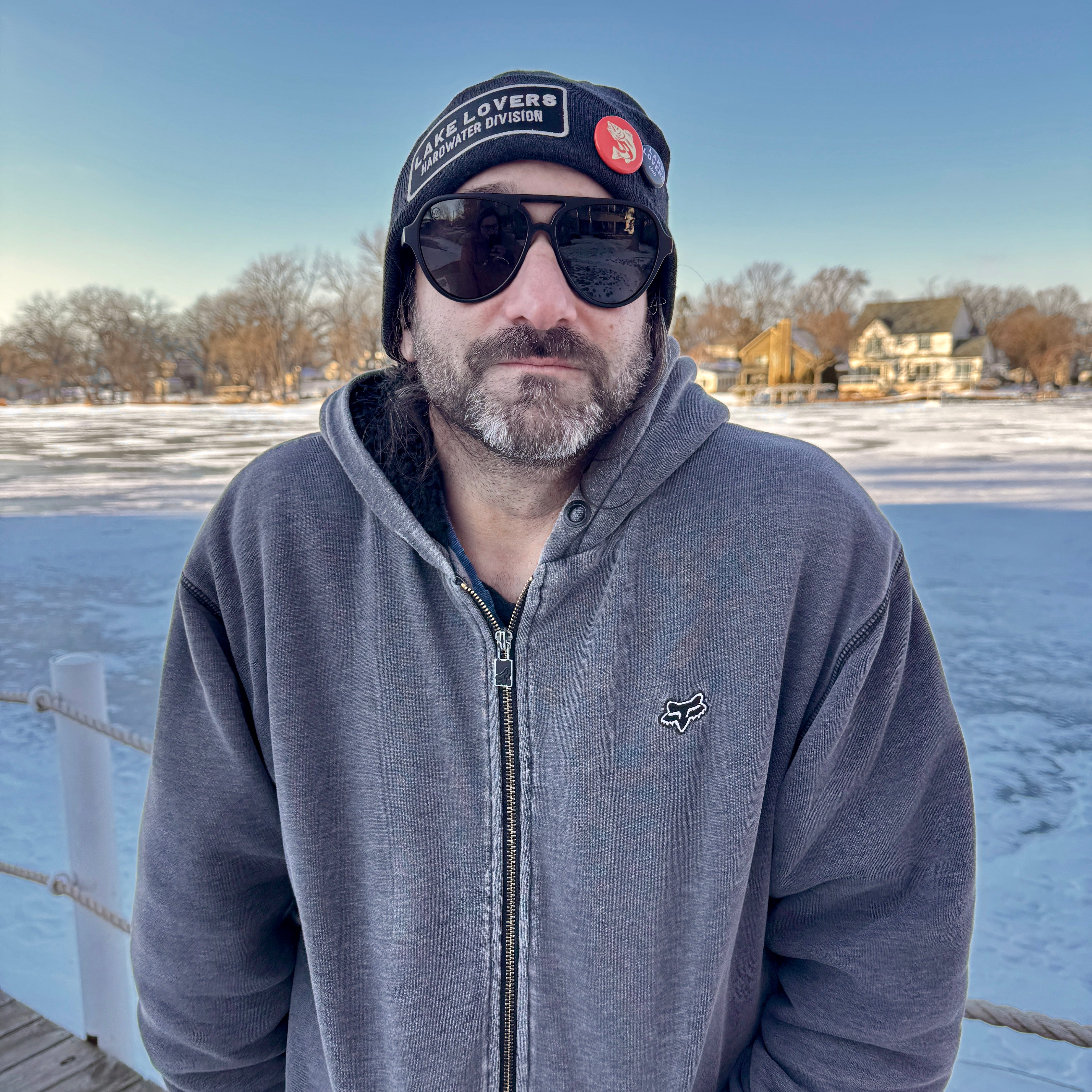 Man wearing sunglasses and a cap standing on a dock with a snowy landscape in the background