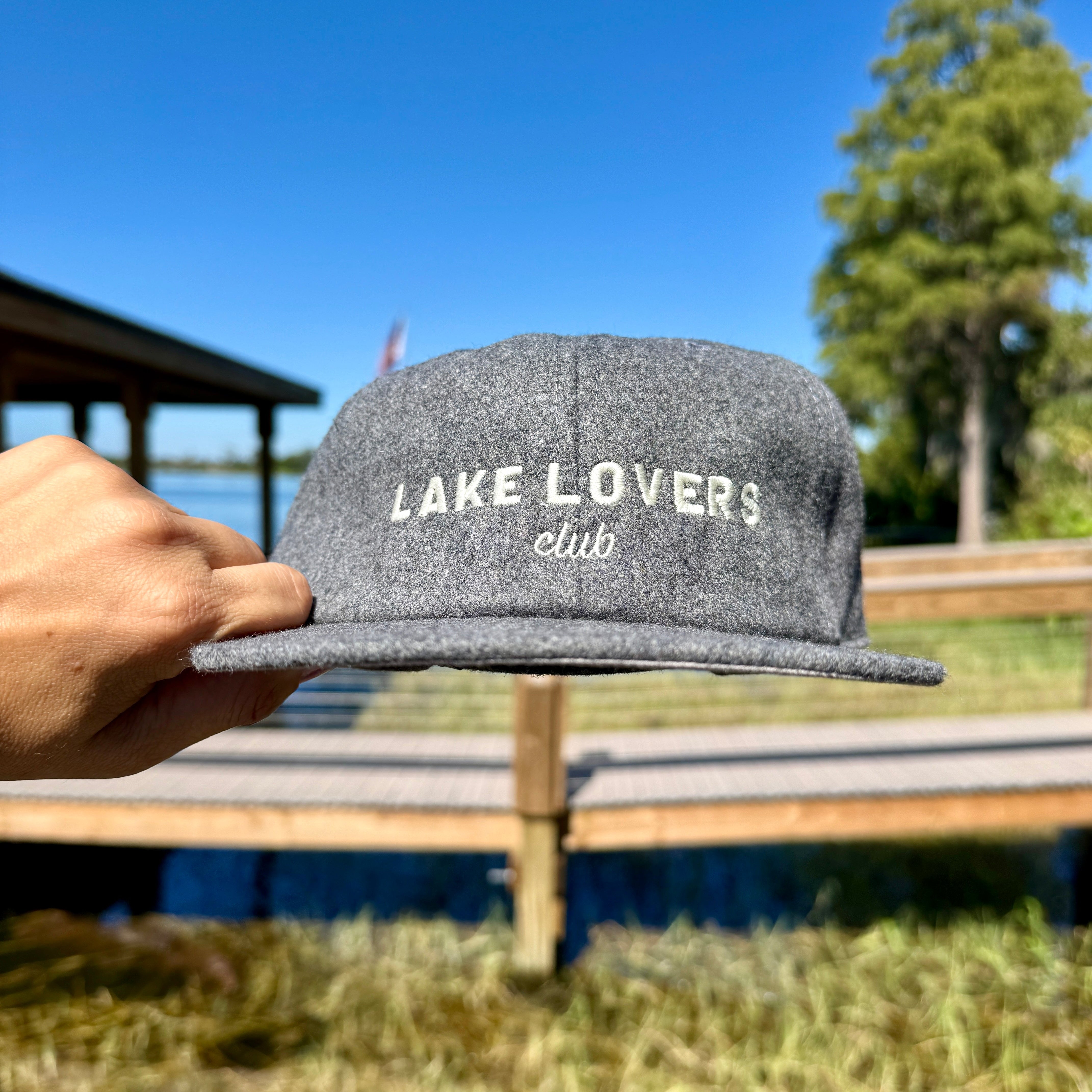 Gray cap with 'Lake Lovers Club' text held by a hand against a lake and trees background