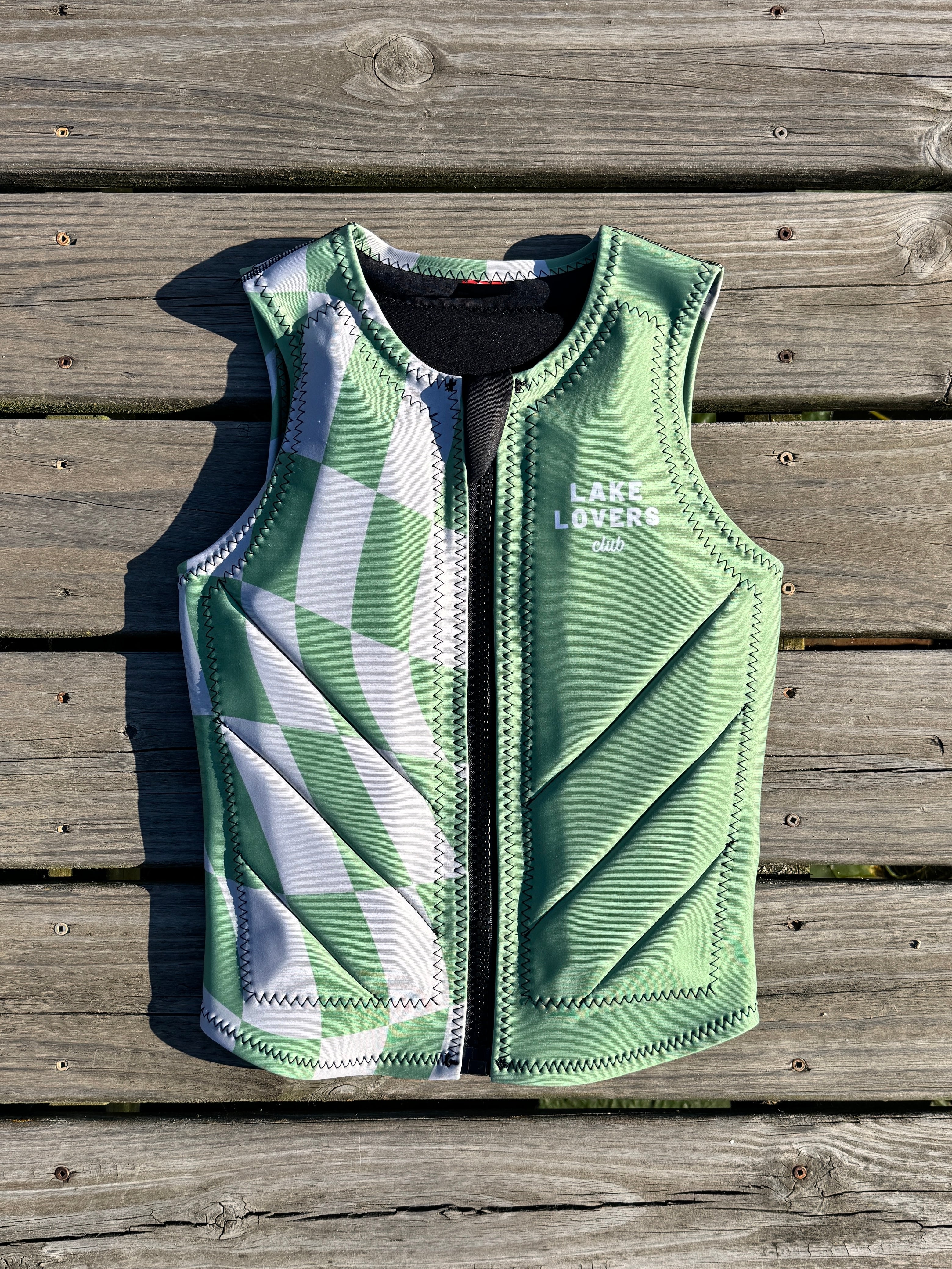 A green and grey patterned zip-up water sports vest laid flat on a weathered wooden plank surface. The vest features a green and grey checkered pattern, a black zipper, and the text 'LAKE LOVERS club' printed on the left chest.