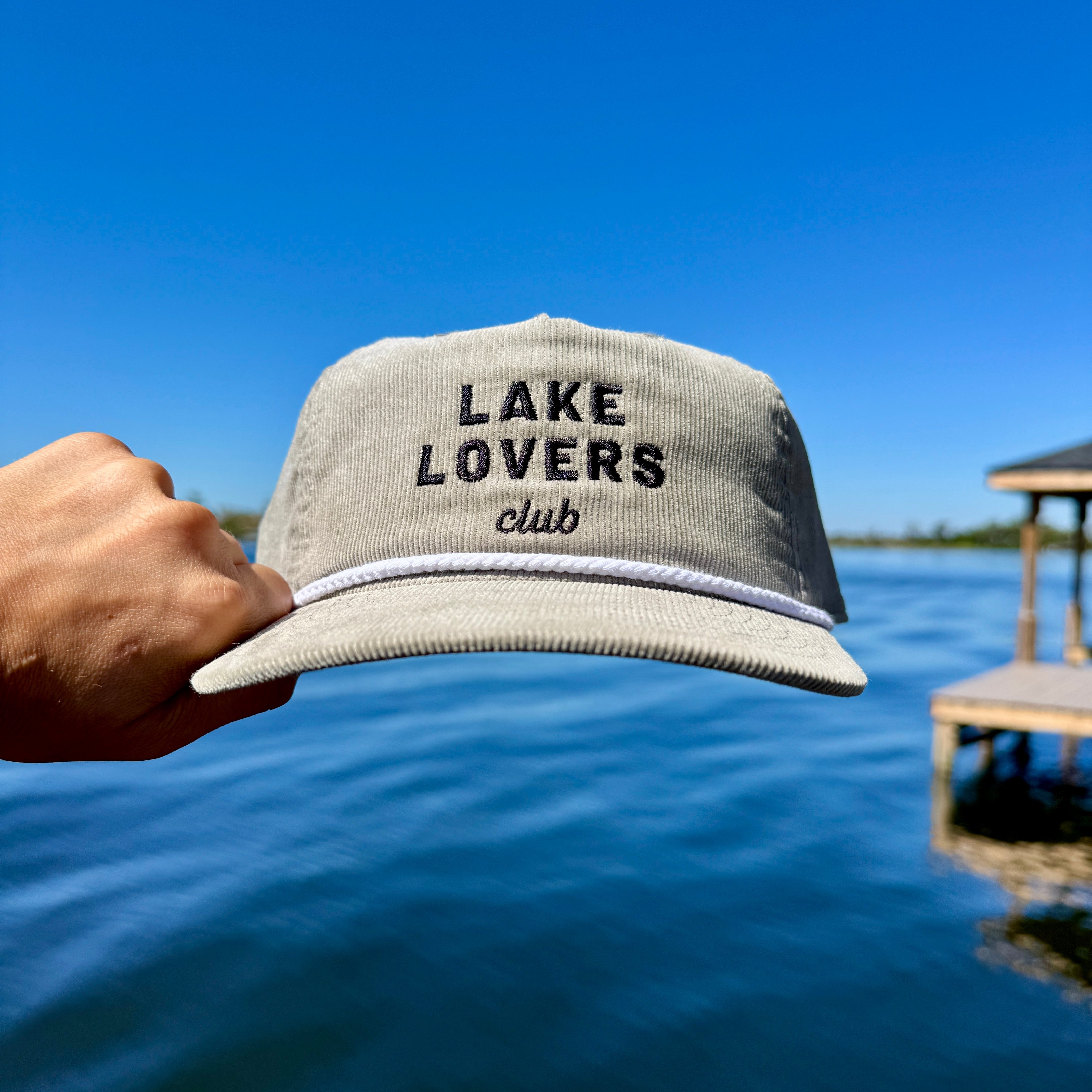 Cap with 'Lake Lovers Club' text held over a lake with a dock in the background.