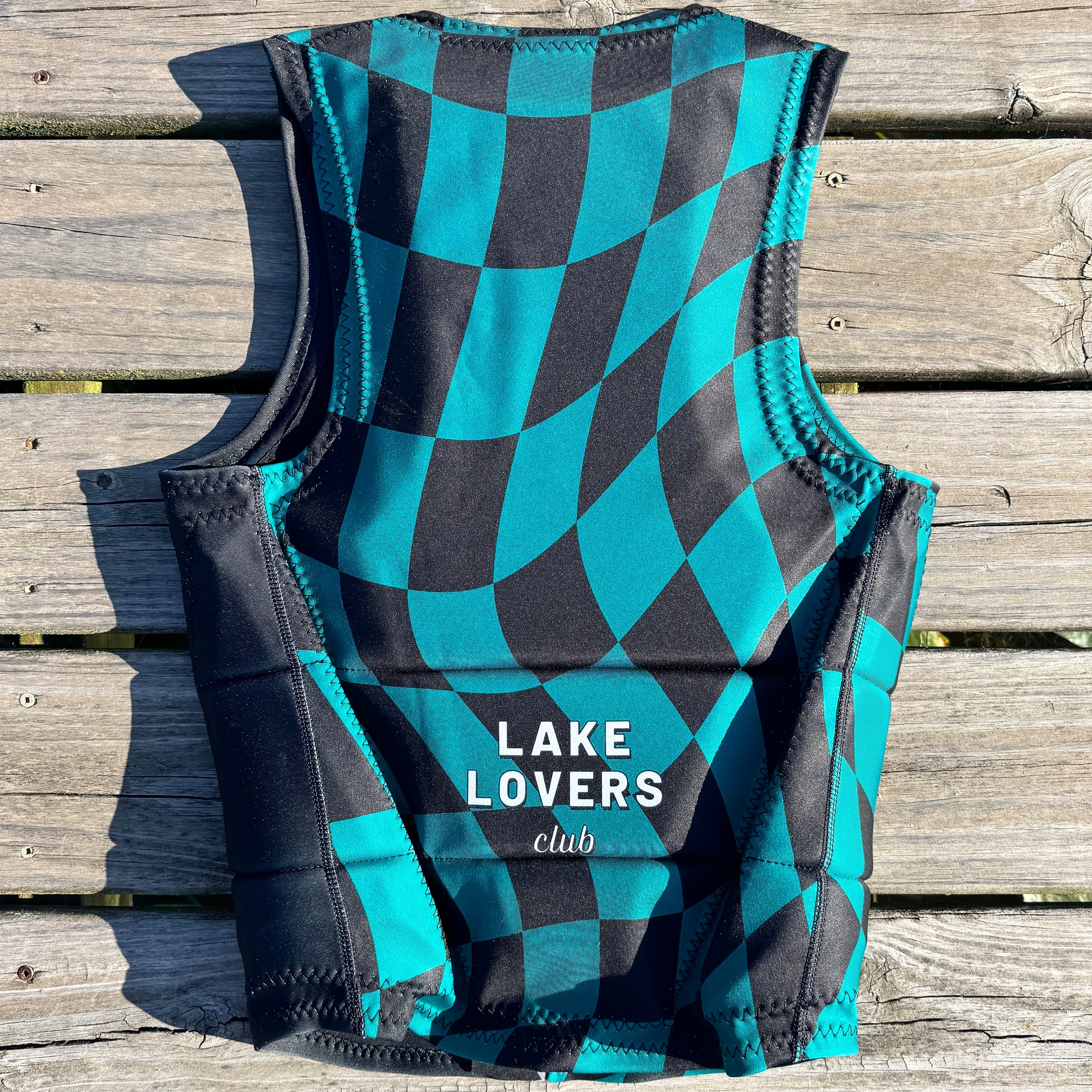 A sleeveless wetsuit vest with a black and teal wavy checkerboard pattern, featuring the text 'LAKE LOVERS club' printed in white on the front. The vest is laid flat on weathered wooden planks.