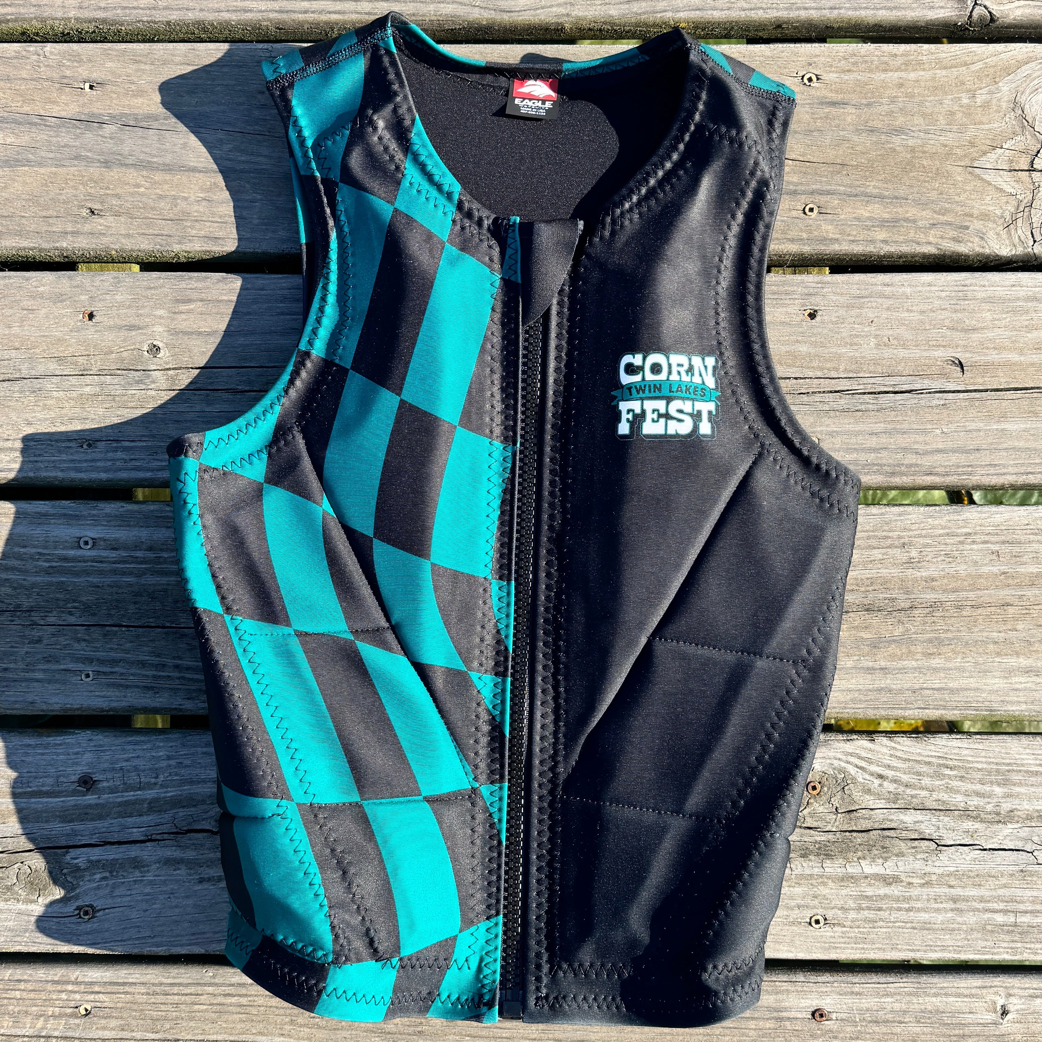 A black and teal checkered water sports vest with a front zipper and 'TWIN LAKES CORN FEST' printed on the chest, laid flat on weathered wooden planks.